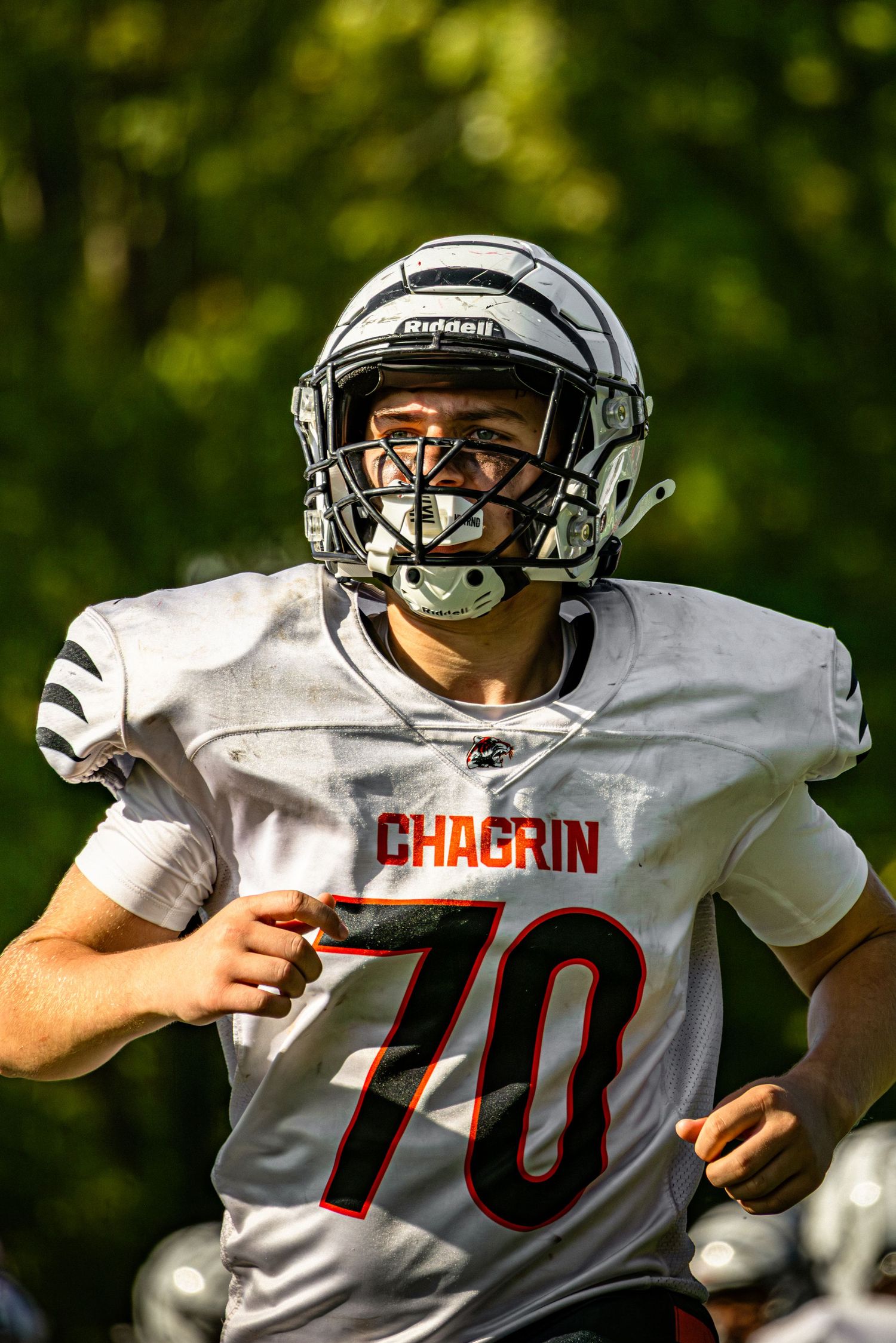 A football player in white Chagrin jersey number 70 runs during a game with green foliage in the background.