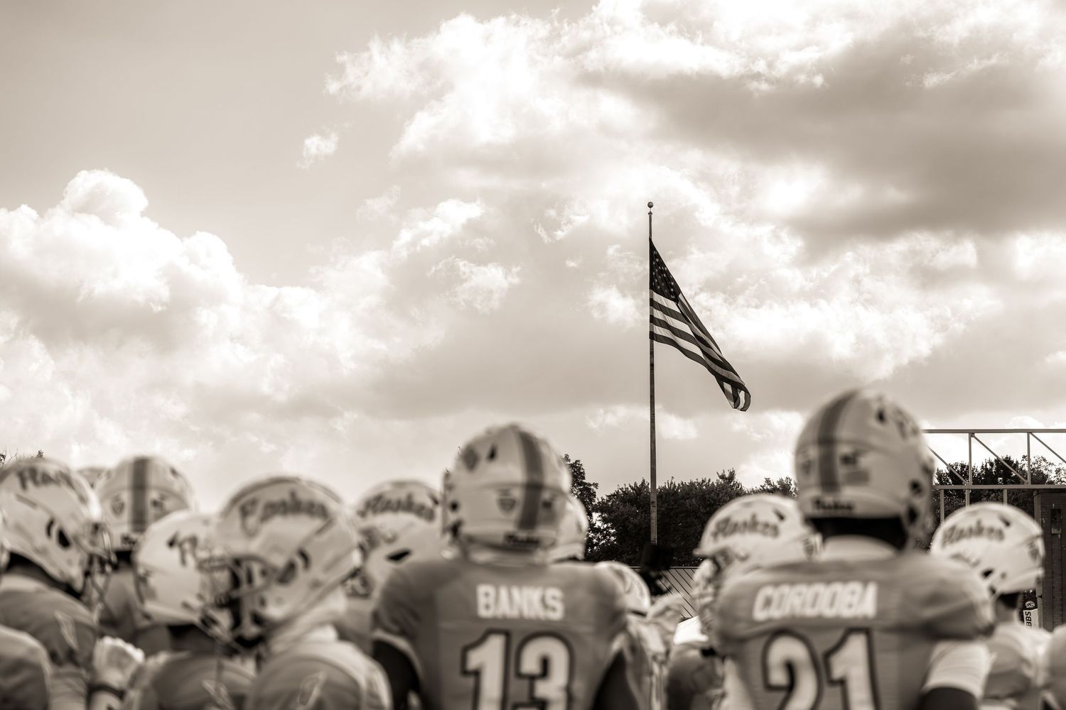 American football players stand on field under dramatic cloudy sky with US flag waving in black and white photograph.
