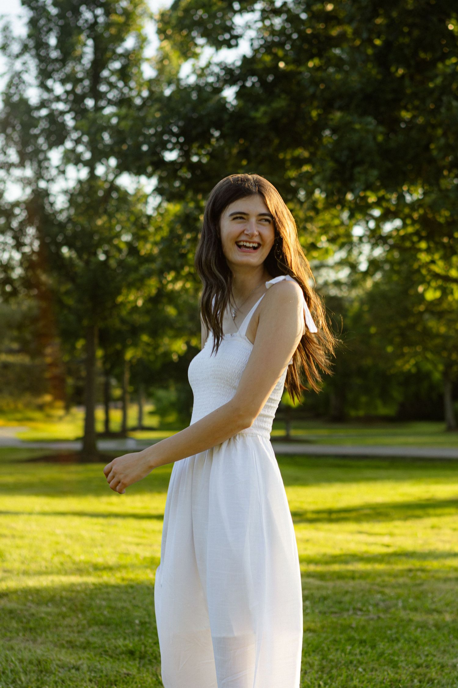 A person in a flowing white summer dress stands in a sunlit park with green trees in the background.