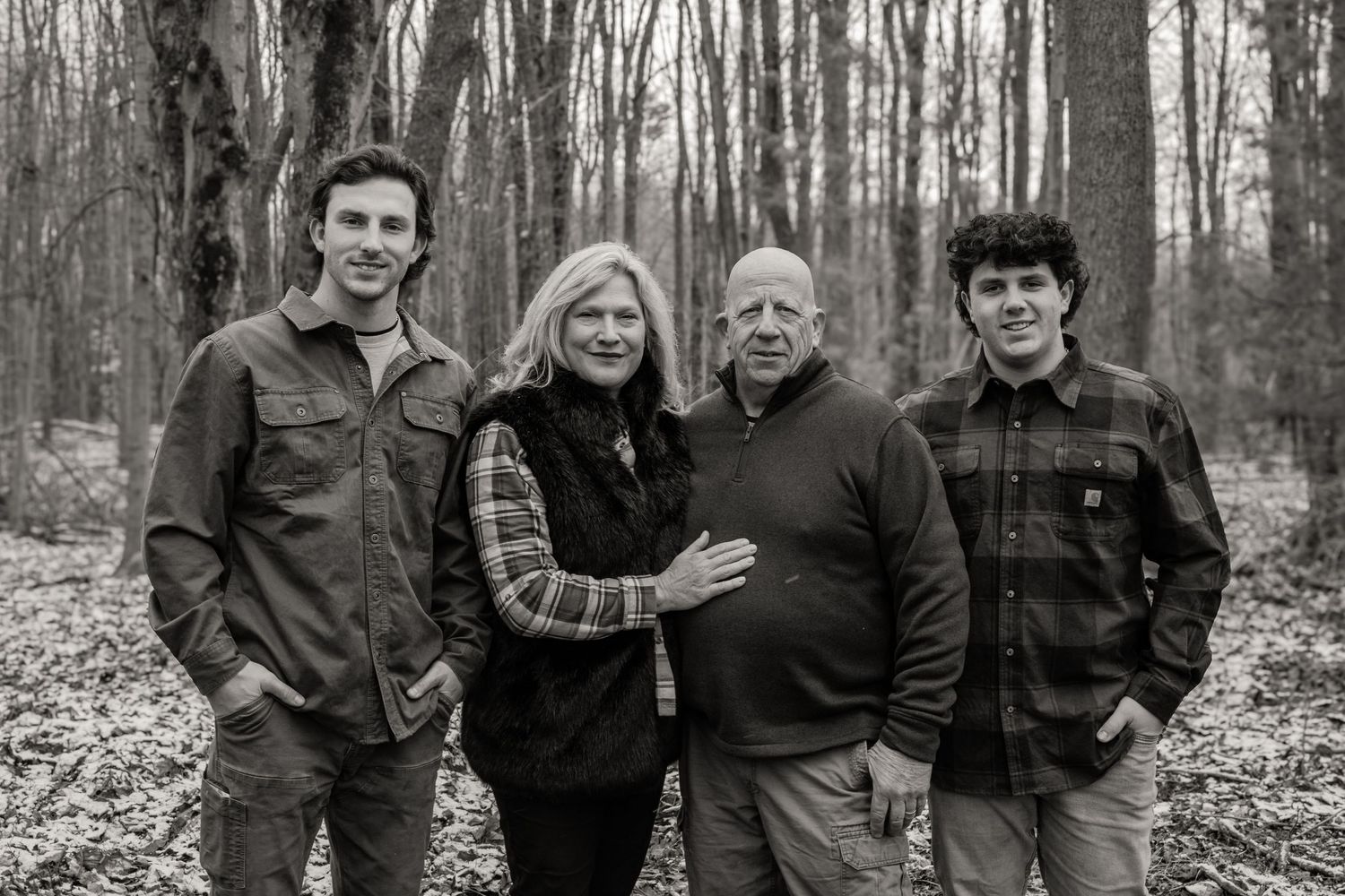 A black and white family portrait of four people standing together in a wooded area during autumn.