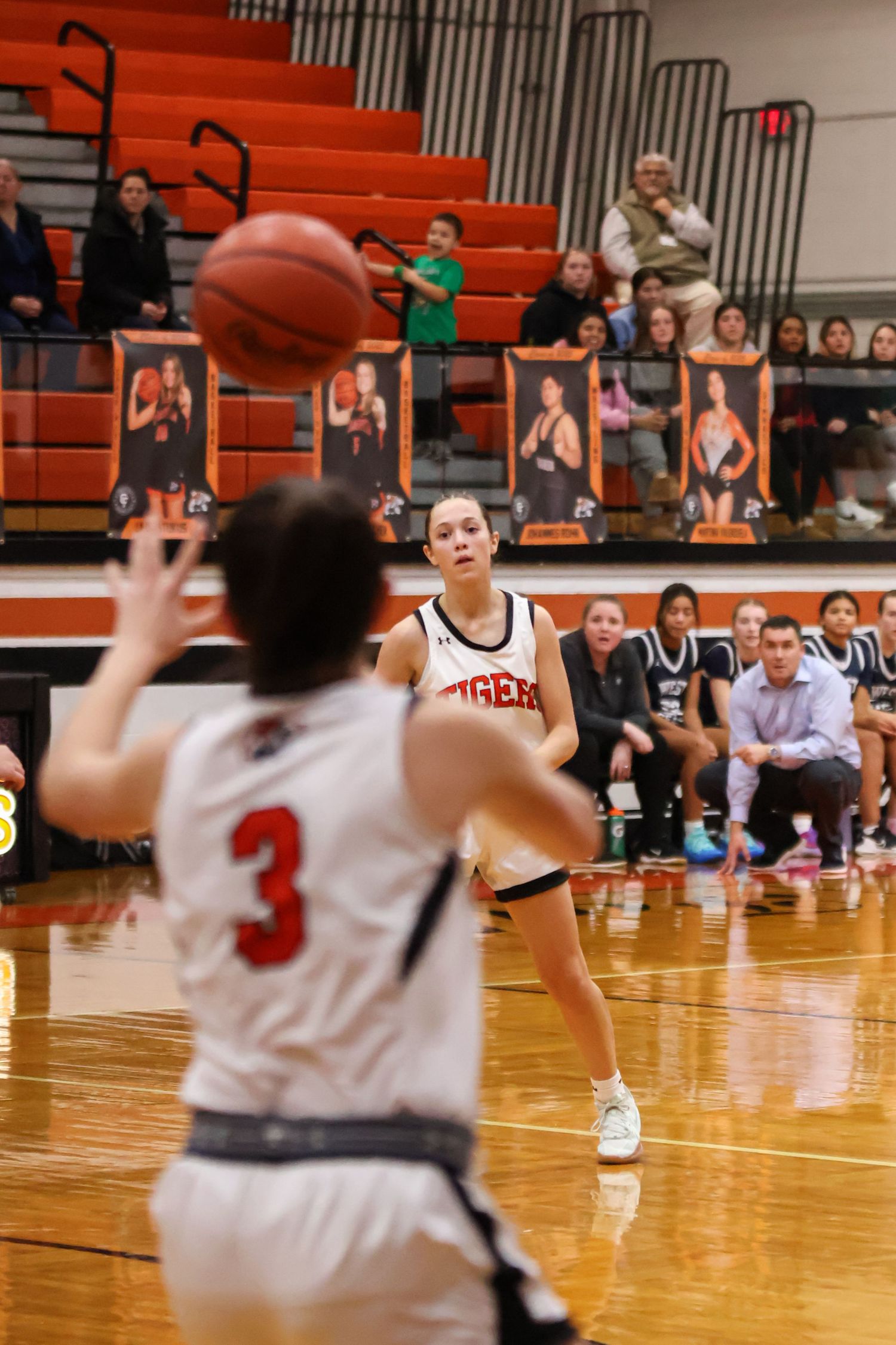 Basketball players reaching for ball on wooden court with orange bleachers in background.