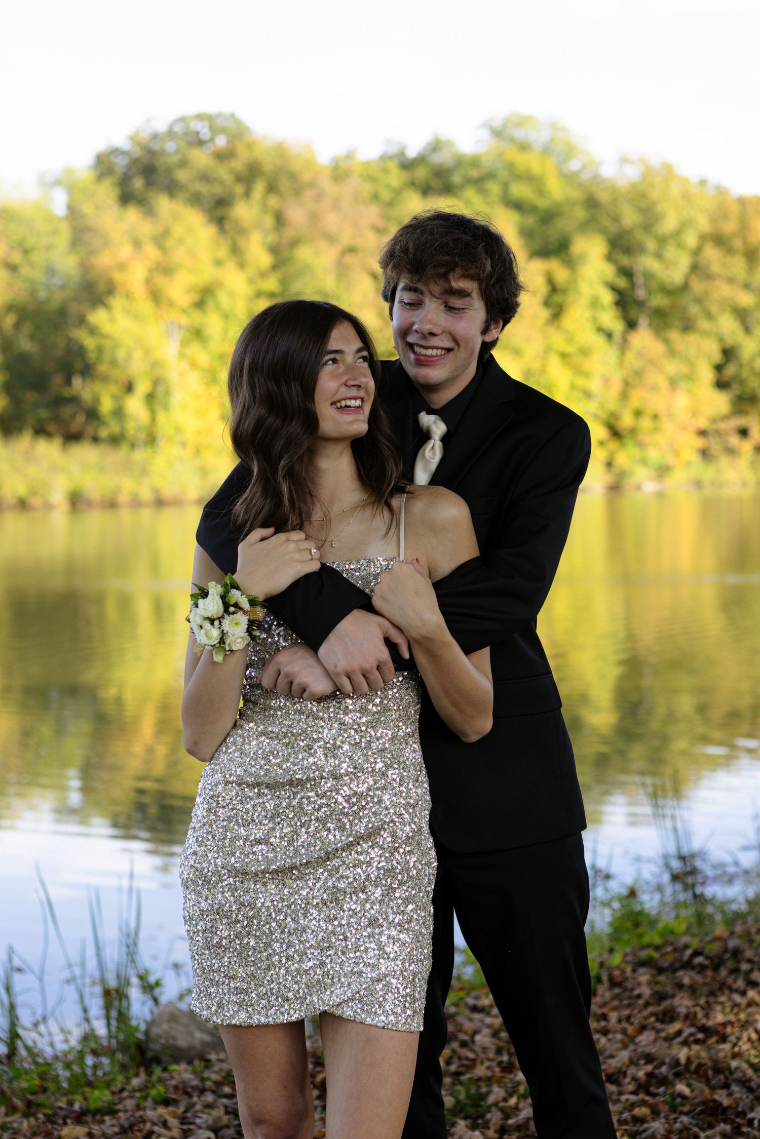 A couple in formal attire shares romantic moments by a serene lake during golden hour in autumn.