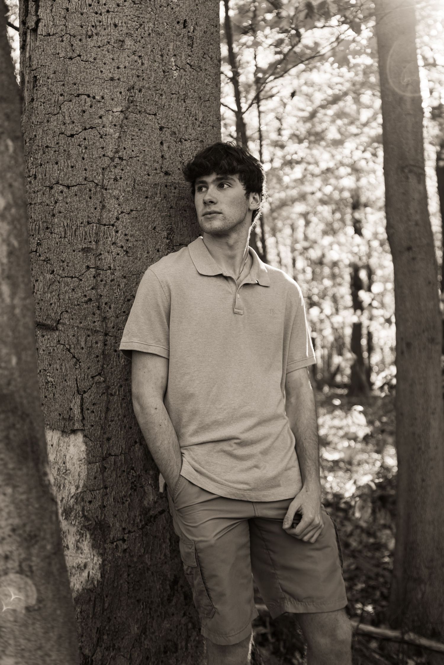 A person in casual attire leans against a large tree trunk in a wooded forest setting.