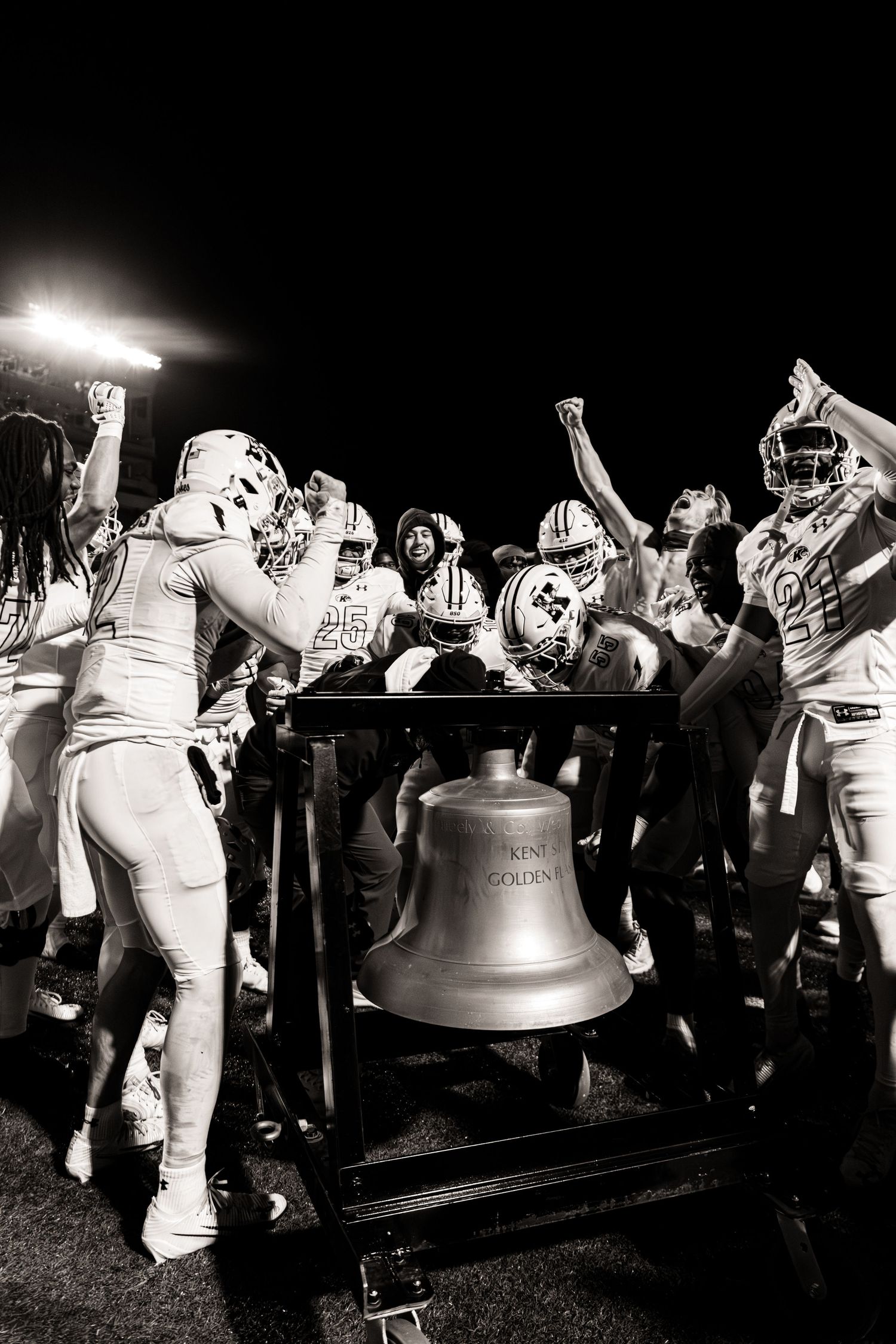 Black and white photo of football players celebrating with the Liberty Bell trophy at a night game.
