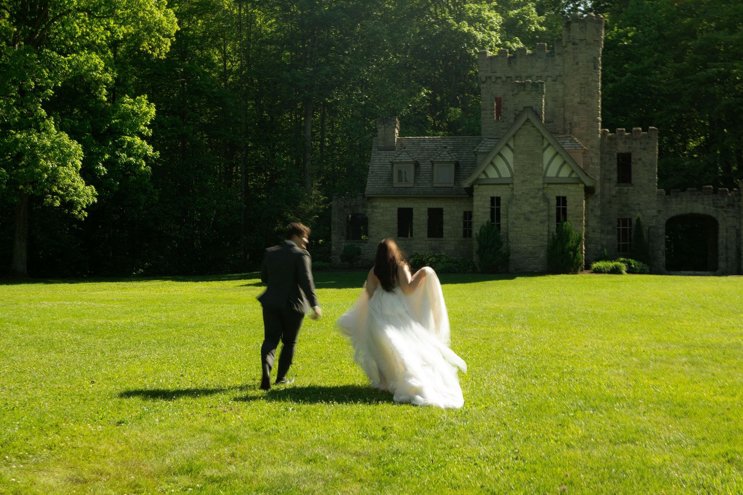 Bride in white gown and groom in black suit running toward a stone castle surrounded by lush green trees.