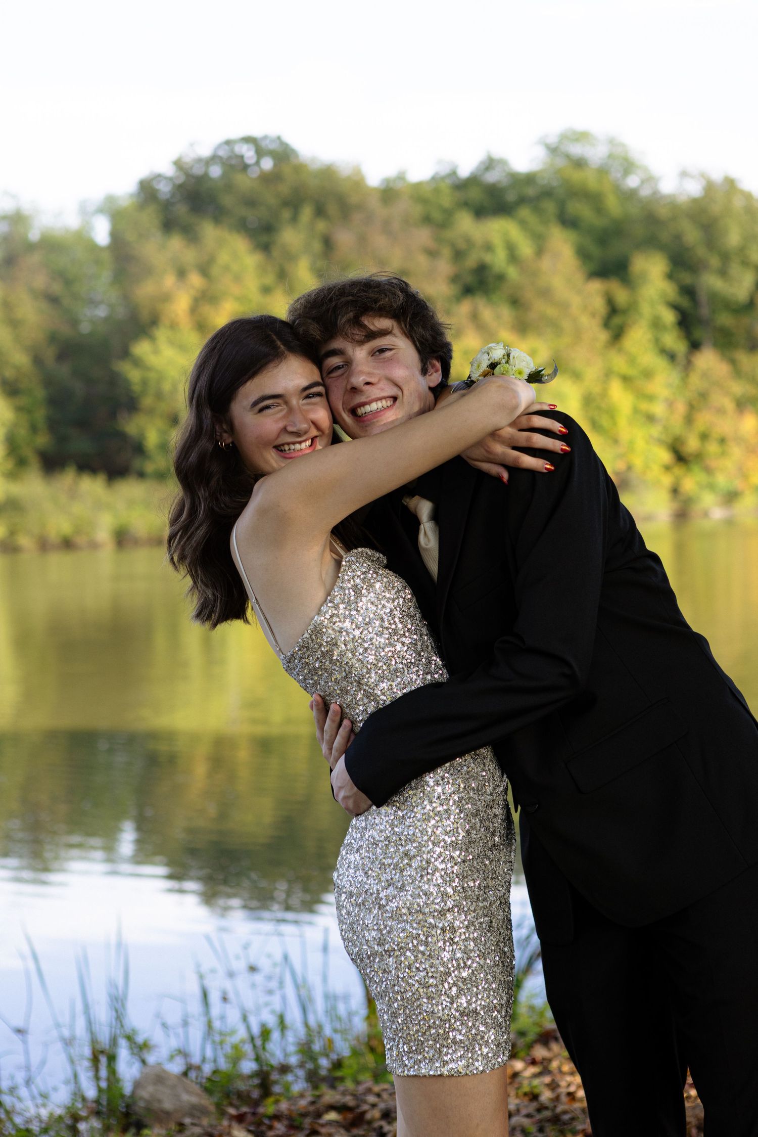 A couple in formal attire embraces and laughs together during a photo session by a scenic lake.