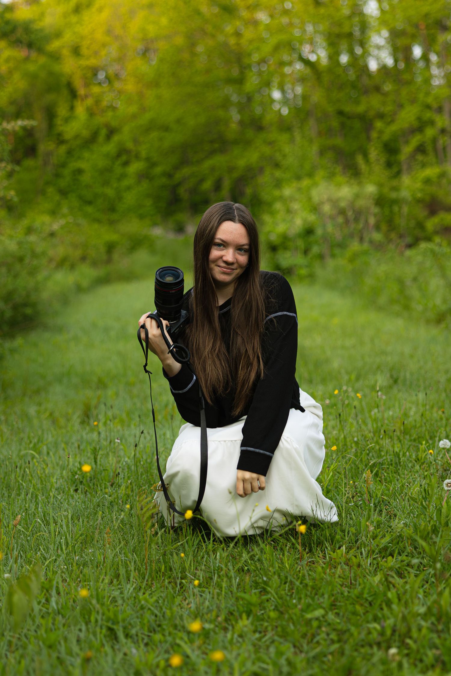 Photographer in black sweater and white skirt kneels with camera in a grassy meadow.