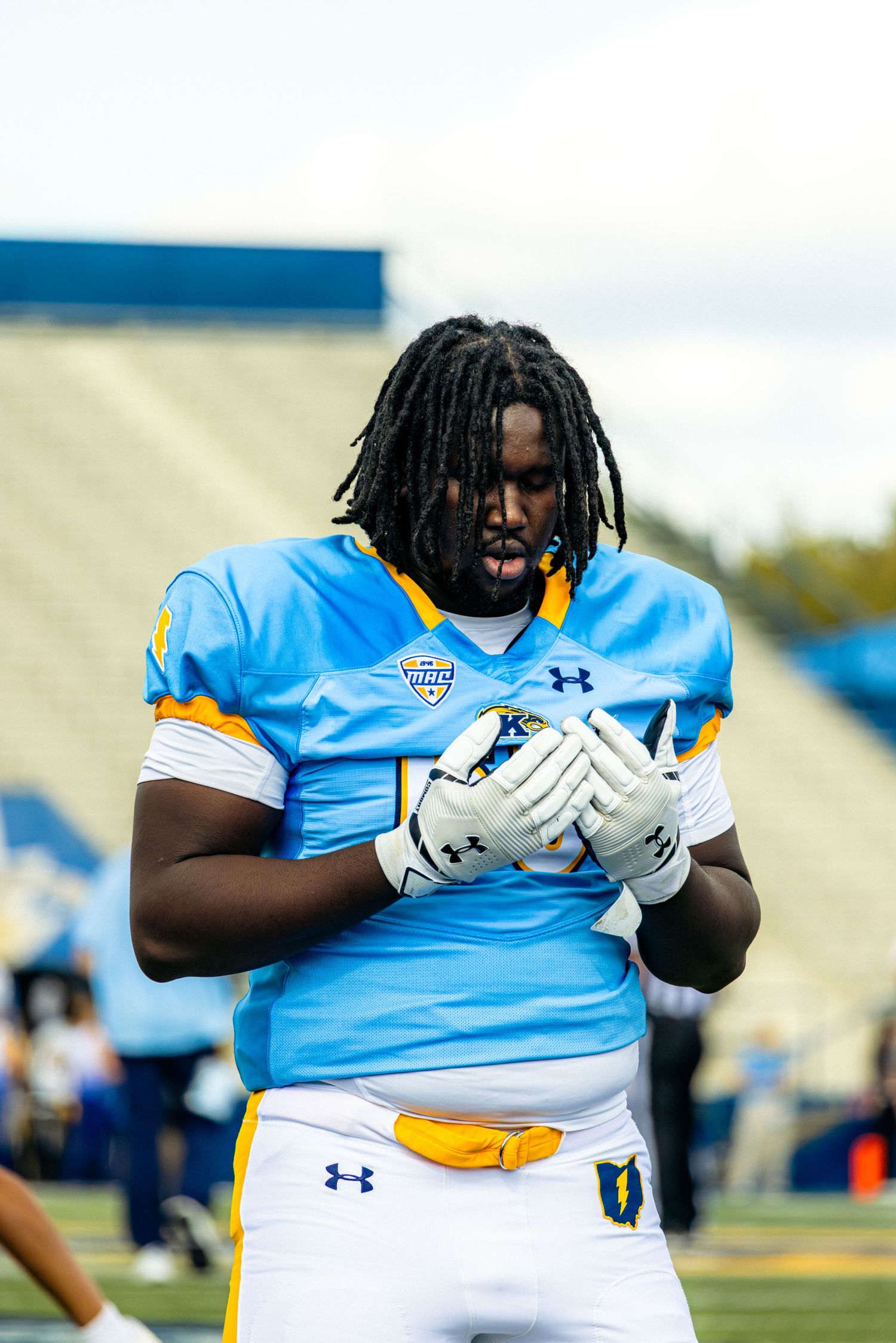 Football player in light blue Under Armour jersey and white gloves stands ready on the field during practice.
