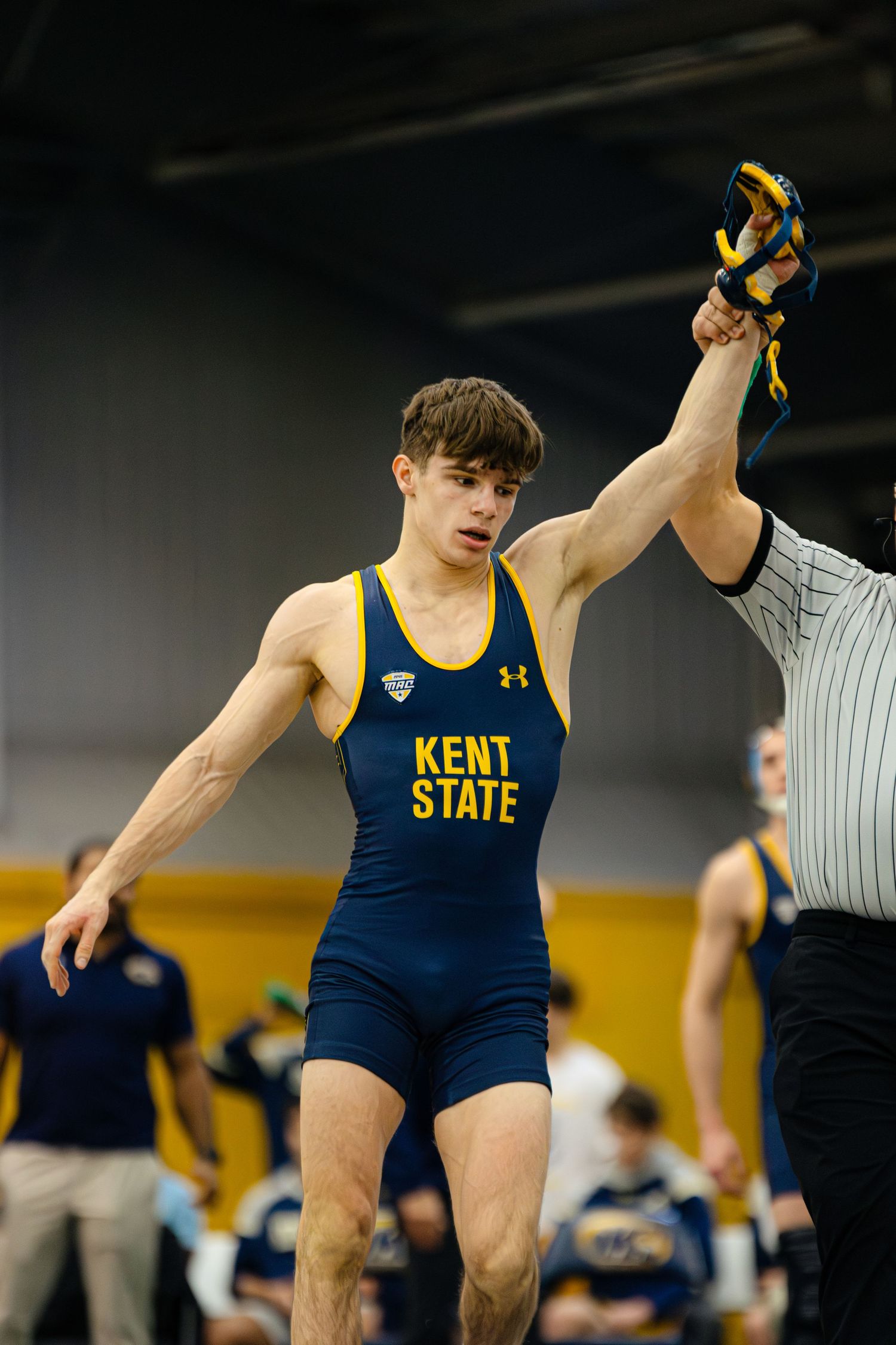 A collegiate wrestler in a navy blue Kent State singlet performs a wrestling move during a match.