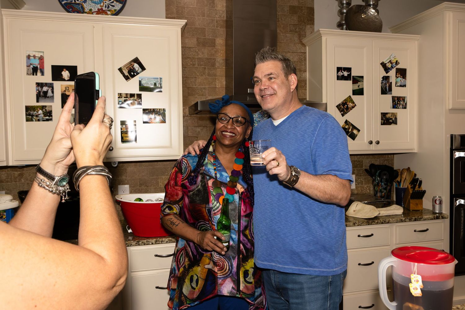 Someone takes a photo of two people posing and smiling together in a kitchen with photographs on the wall behind them.