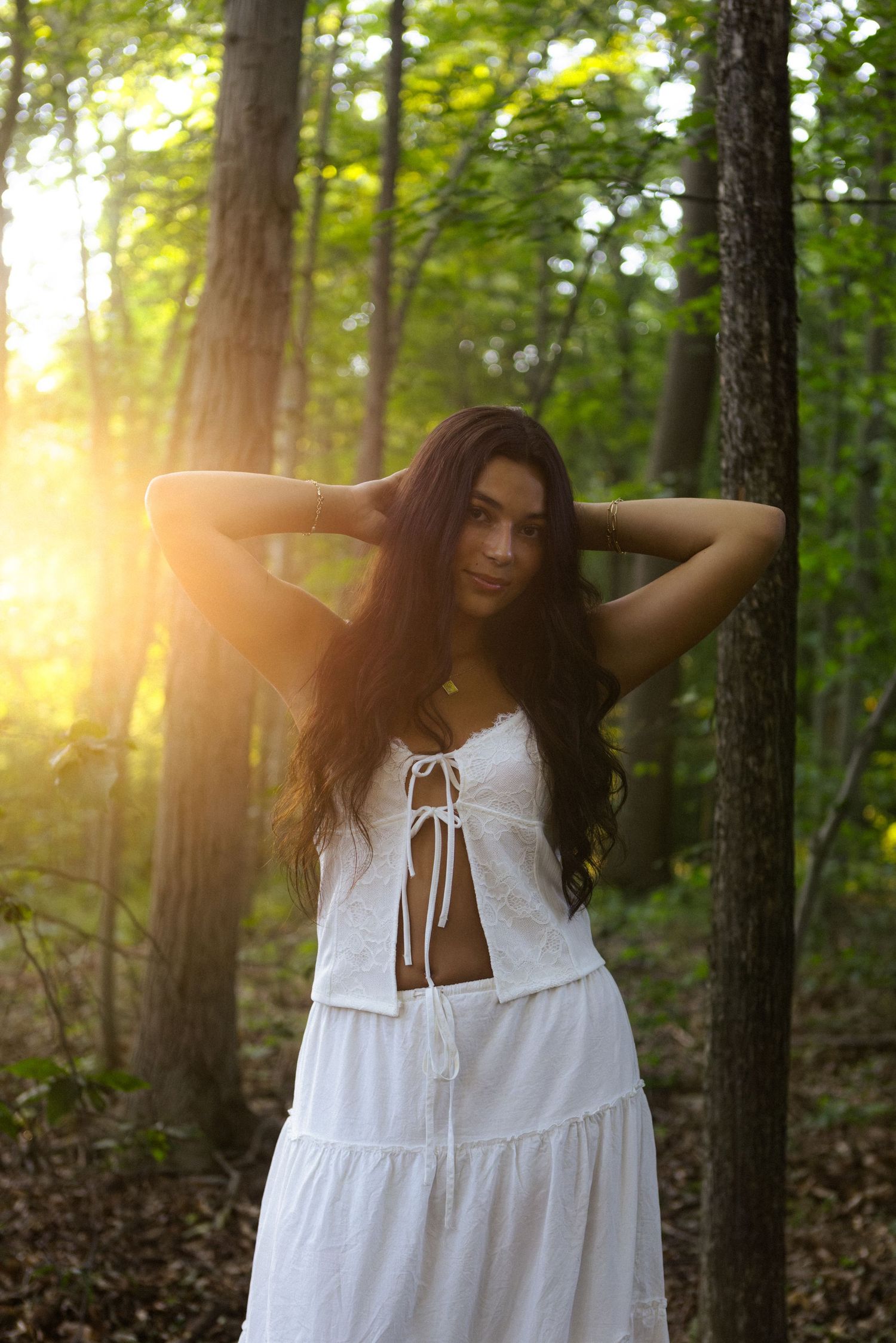 A woman in a white sundress poses gracefully in a sunlit forest setting with golden light filtering through trees.