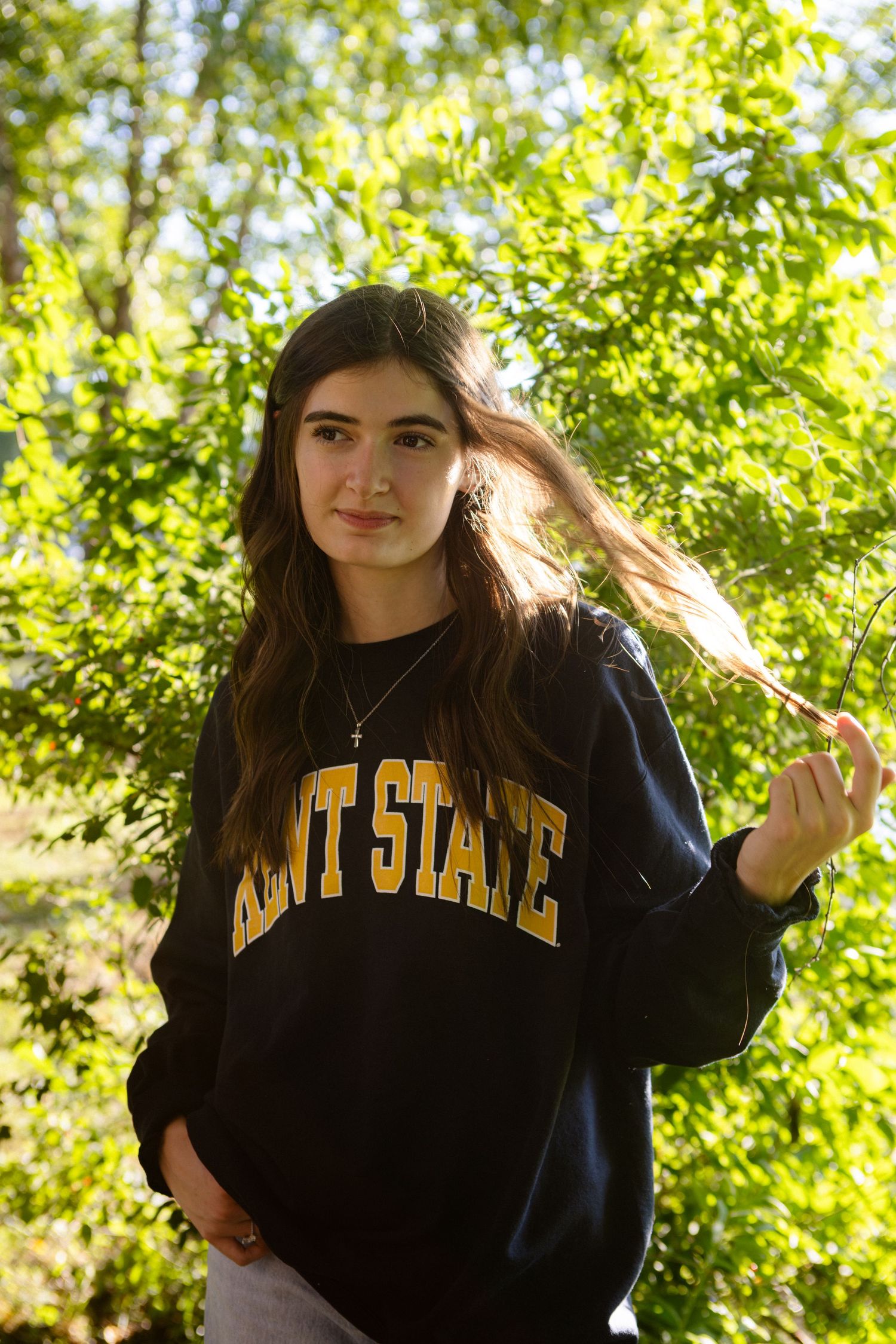 Person wearing a navy blue Kent State sweatshirt outdoors with sunlit foliage in the background.