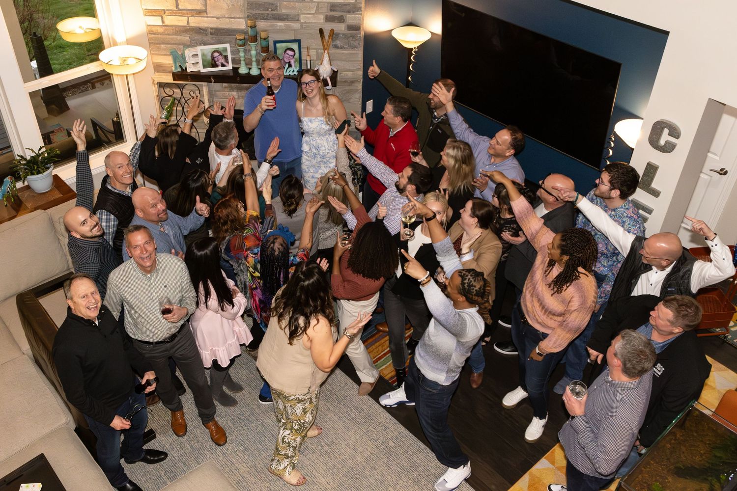 Large group of party attendees gathered in a home living room with raised hands celebrating together.