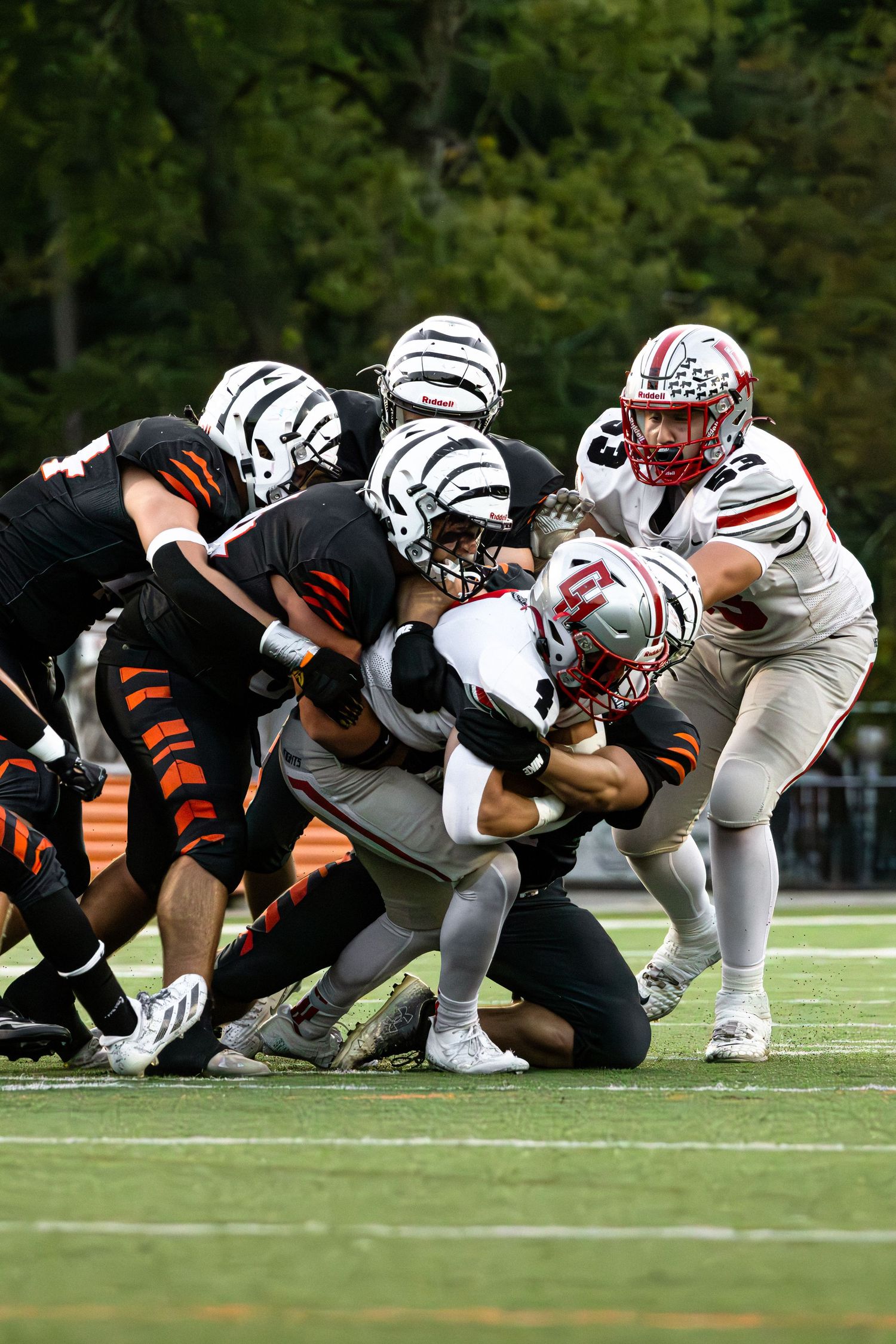 Football players in black and white uniforms clash during a tackle play on the field with trees in background.
