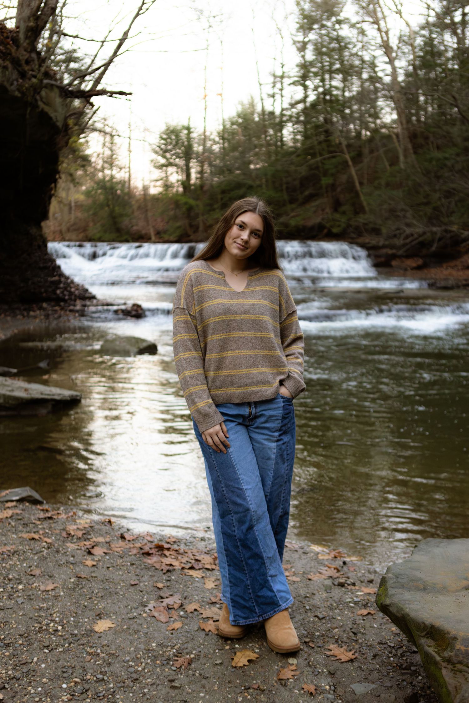 Someone in a striped sweater and jeans poses near a cascading waterfall in a wooded setting during autumn.