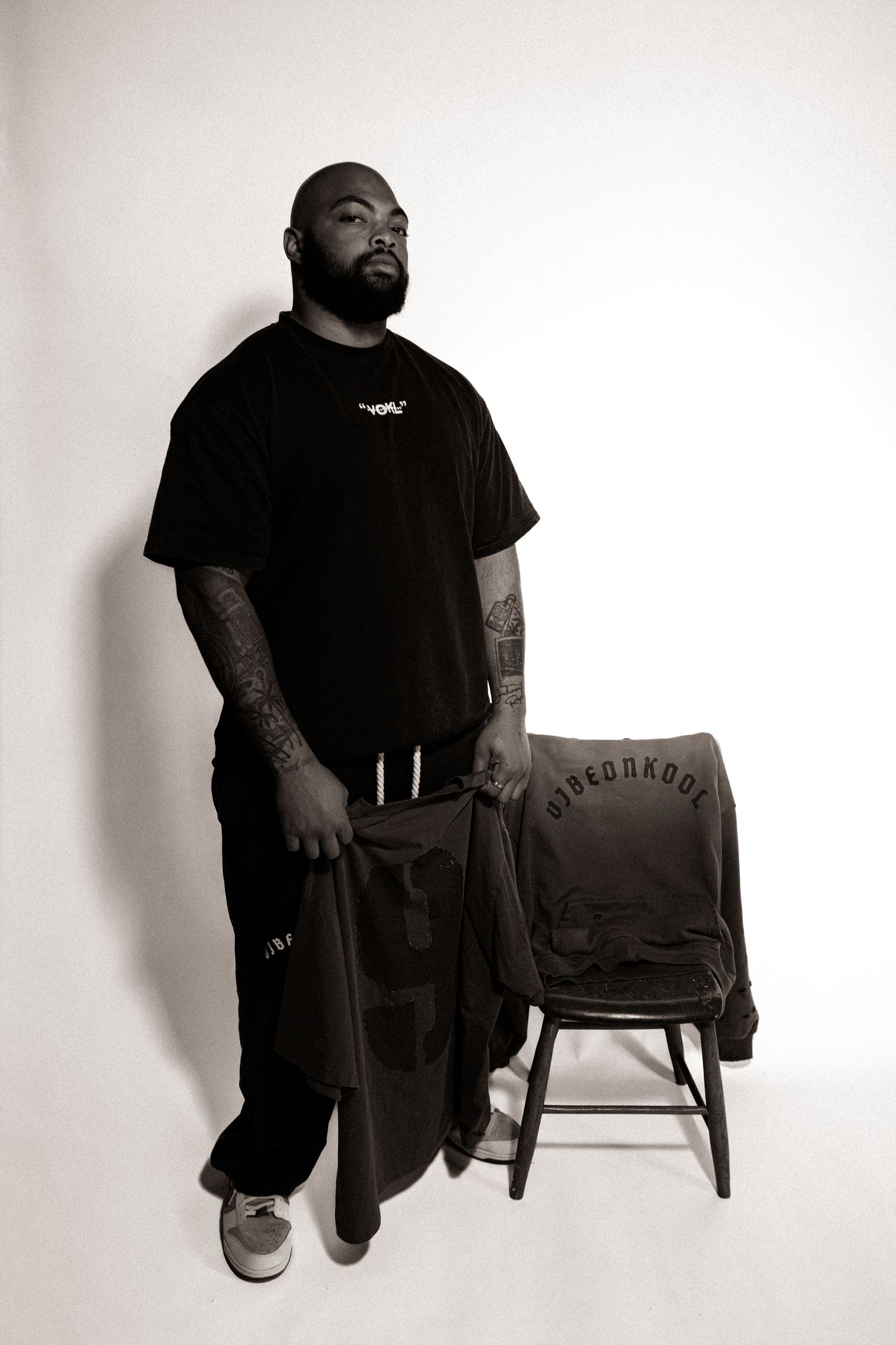 Tattooed man in black outfit stands beside a stool with a branded hoodie in a studio portrait.