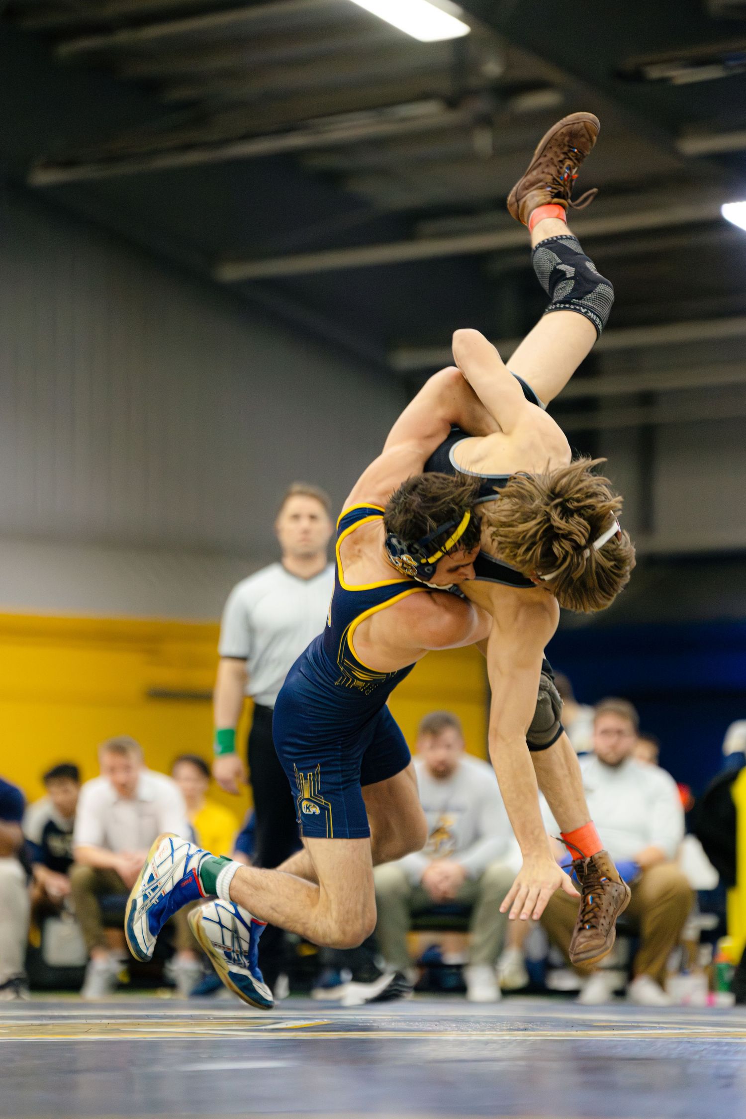 A sequence of wrestling moves shows a takedown attempt on a blue mat during an indoor match.