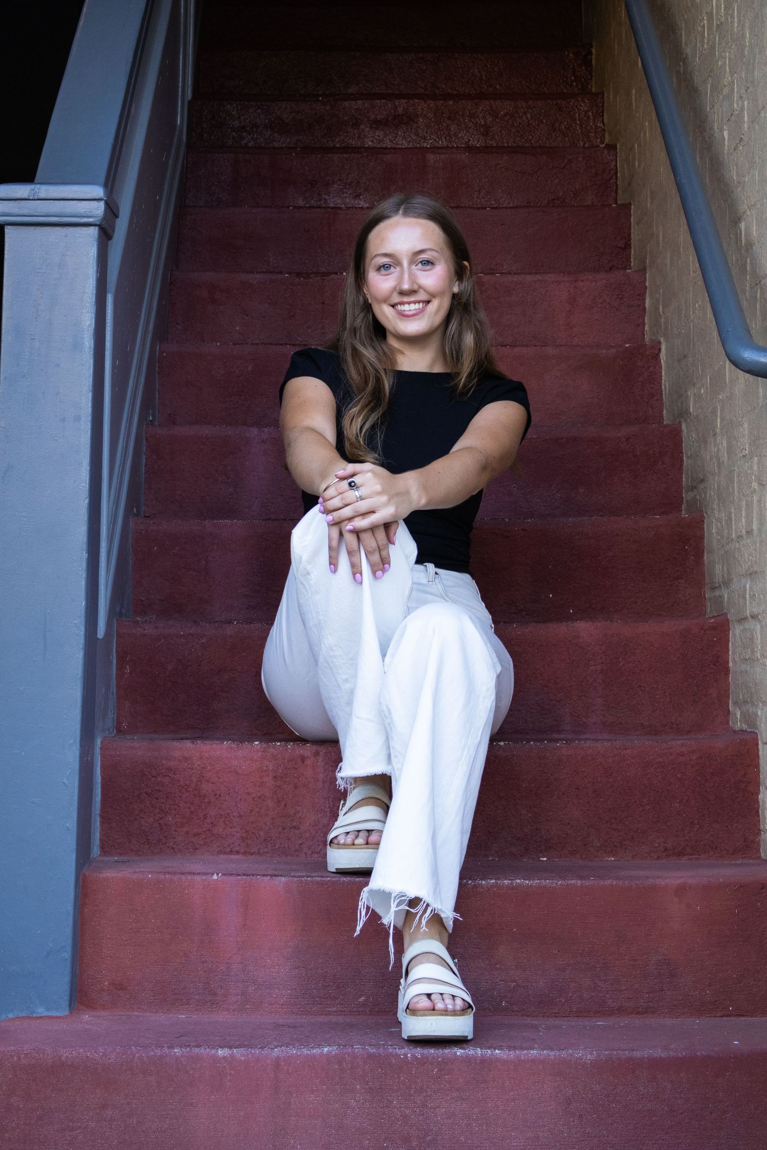 A person sitting on burgundy painted wooden stairs in casual attire smiling at the camera.
