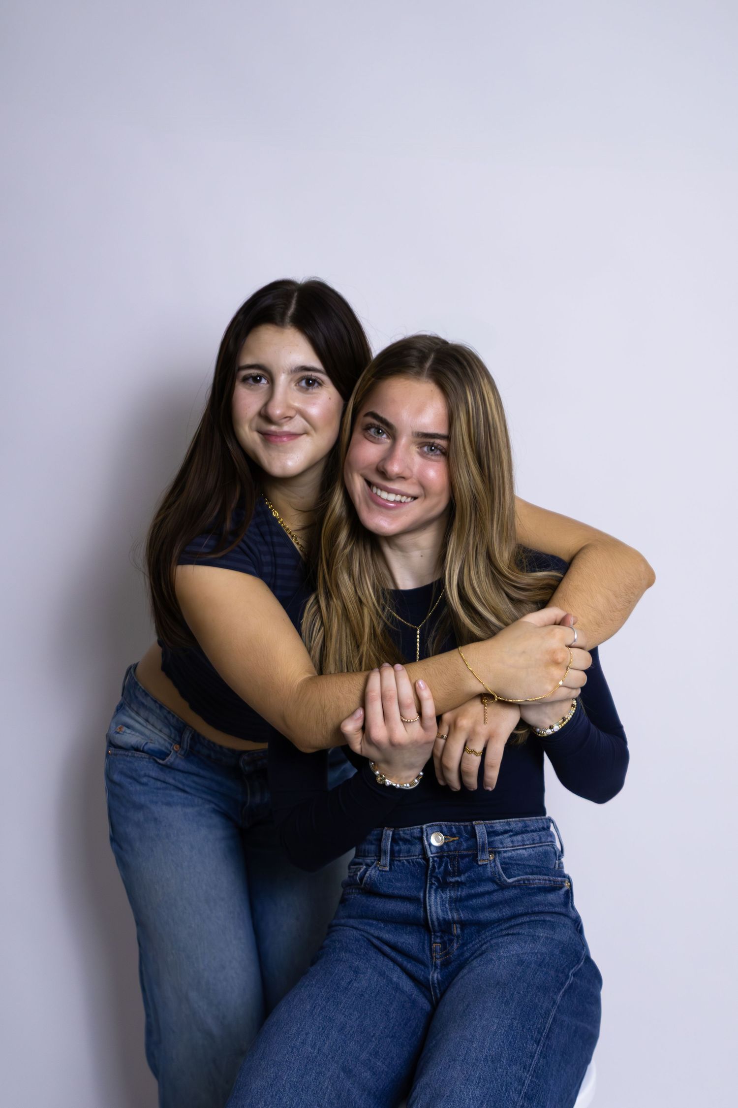 Two young women in jeans and dark tops embrace while posing against a light gray wall.
