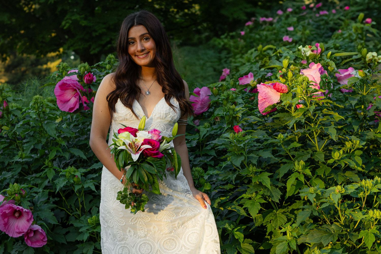 A bride in a white lace wedding dress holds a colorful bouquet while standing in a garden of pink roses.