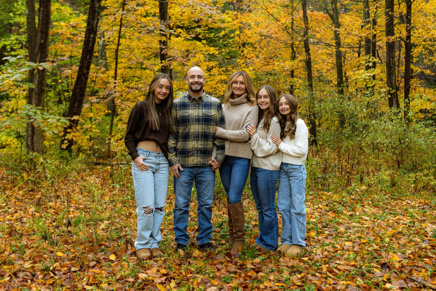 A group stands together on fallen autumn leaves in a forest with vibrant yellow foliage in the background.