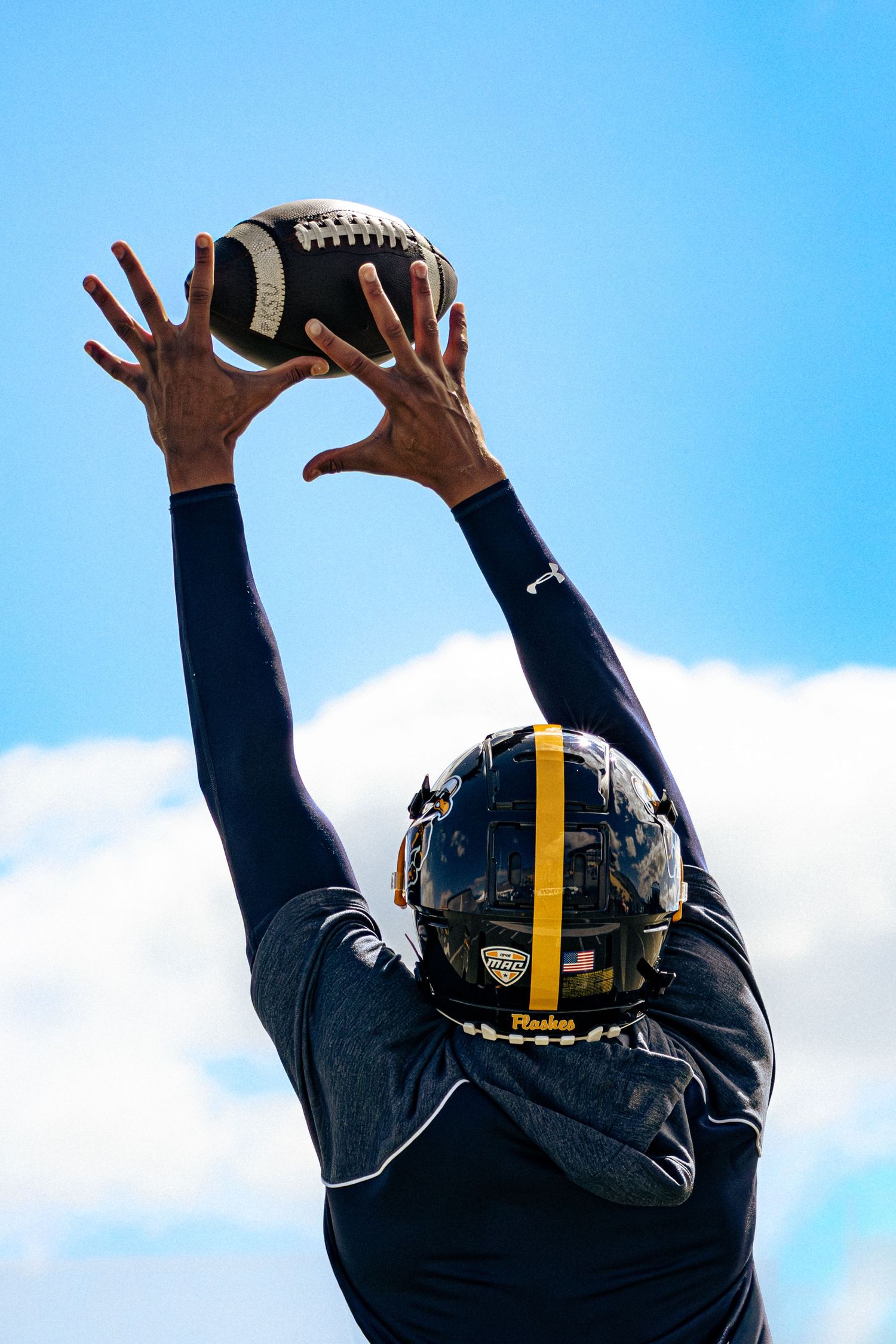 Person in black Pittsburgh Steelers jersey raises hands to catch a football against blue sky with white clouds.
