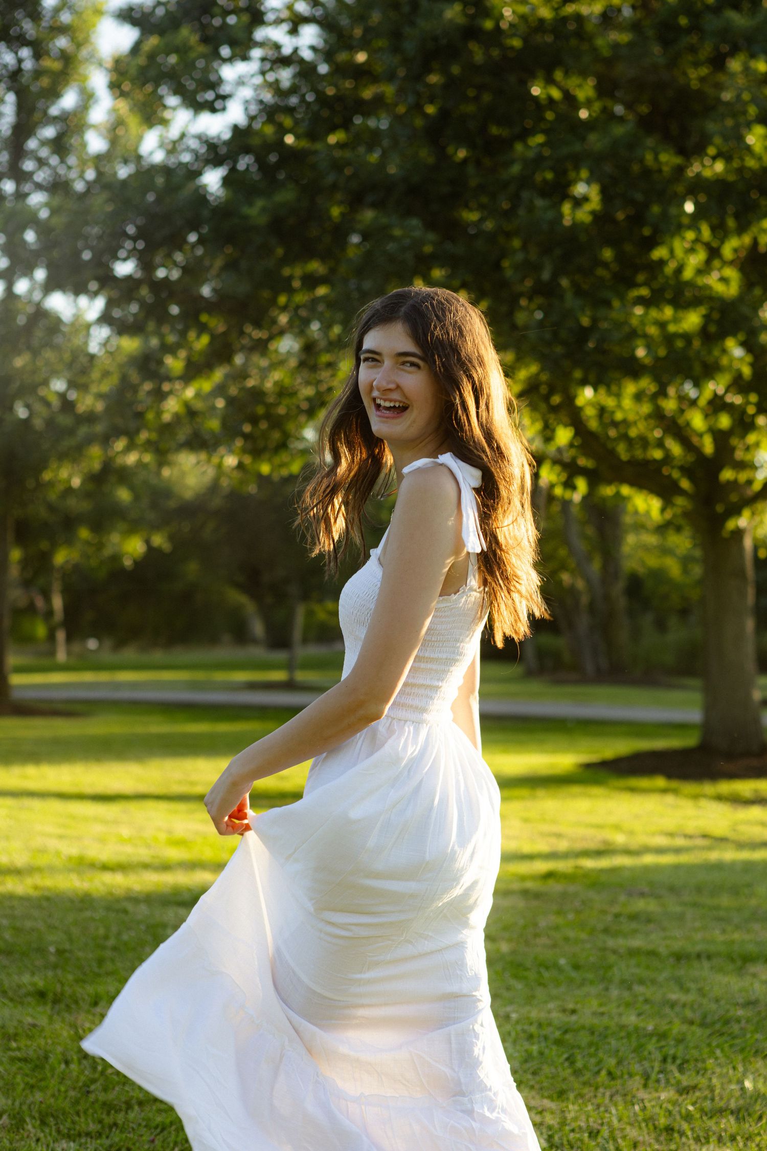 A figure in a flowing white dress twirls gracefully in a sunlit park setting during golden hour.