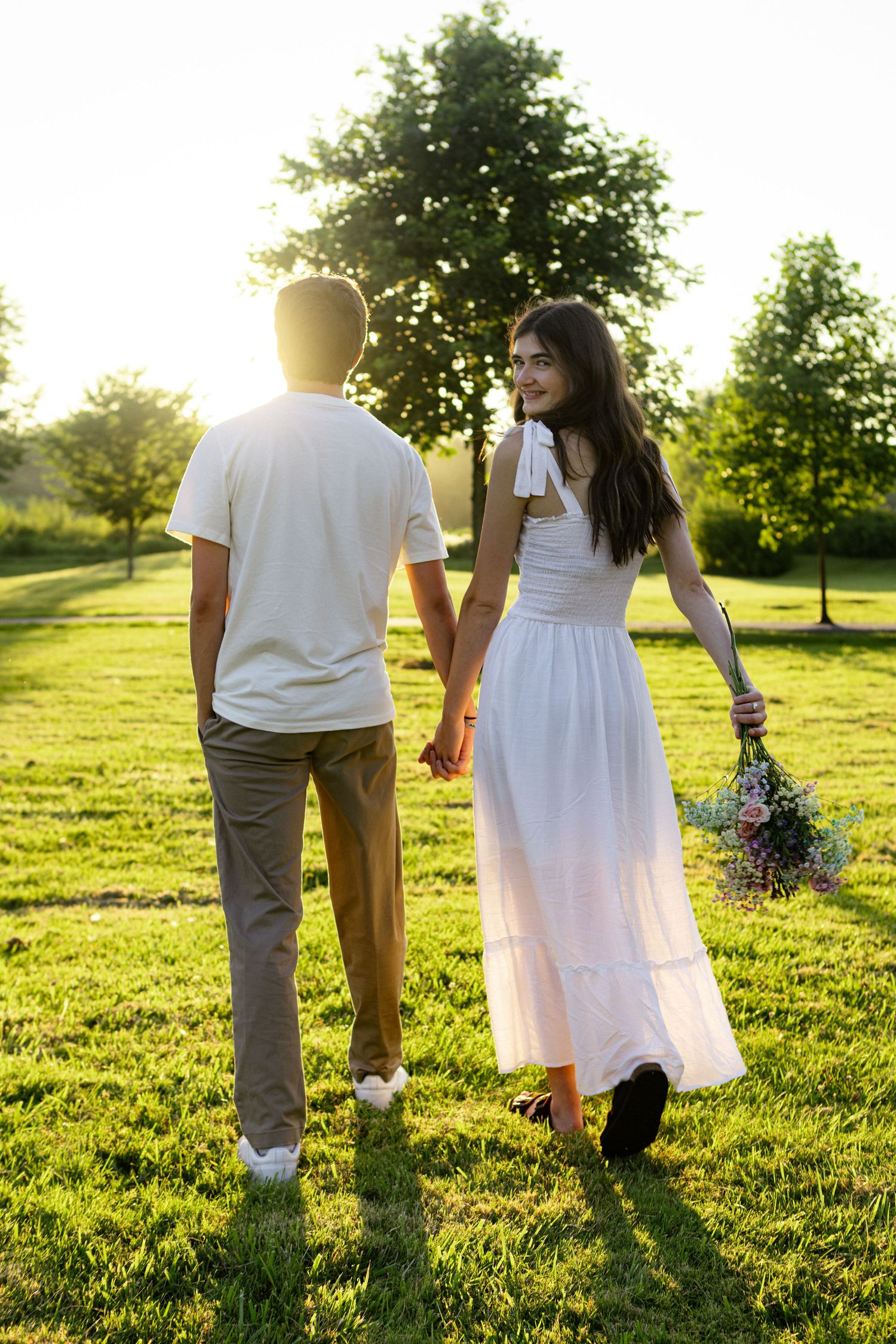 A couple walks hand in hand through a sunlit grassy field on a warm summer evening.