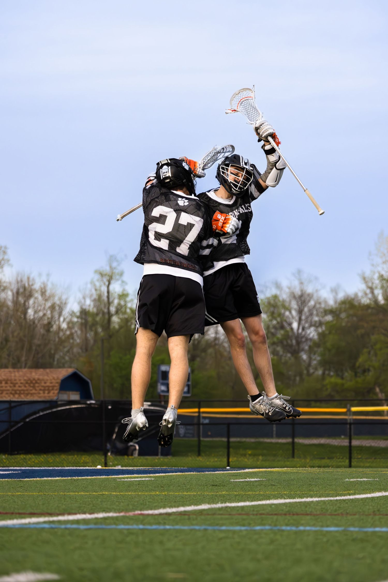 Two lacrosse players leap high to celebrate during game on turf field.