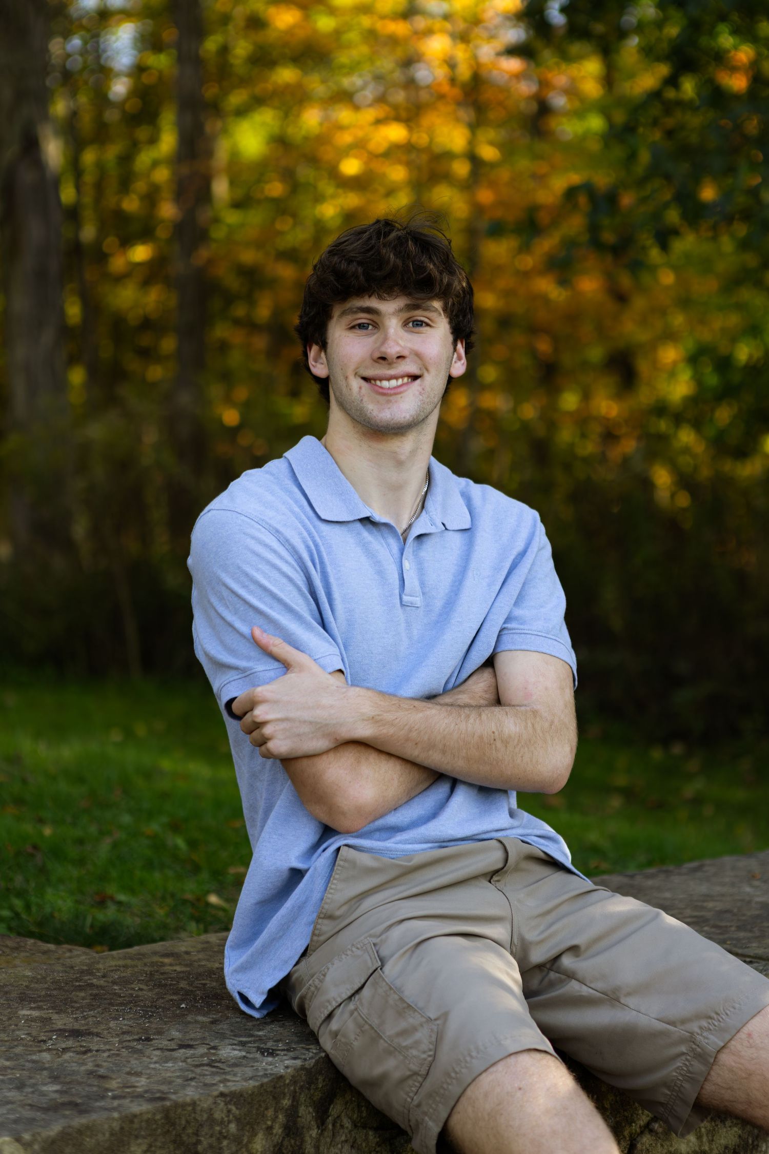 A student in light blue polo and khaki shorts poses casually outdoors against an autumn forest backdrop.