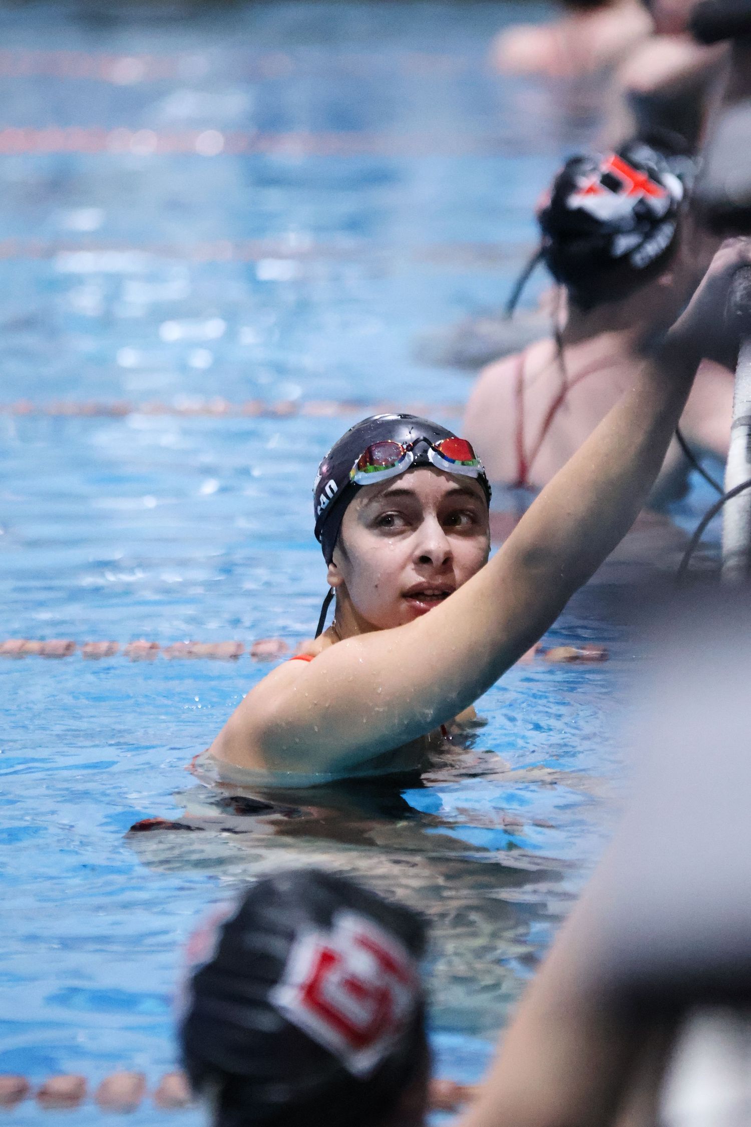 Swimmer in black cap and goggles holds onto pool wall during competition.