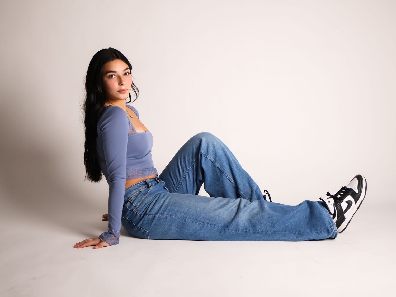Person in casual denim outfit lying sideways on studio floor with white backdrop in relaxed pose.