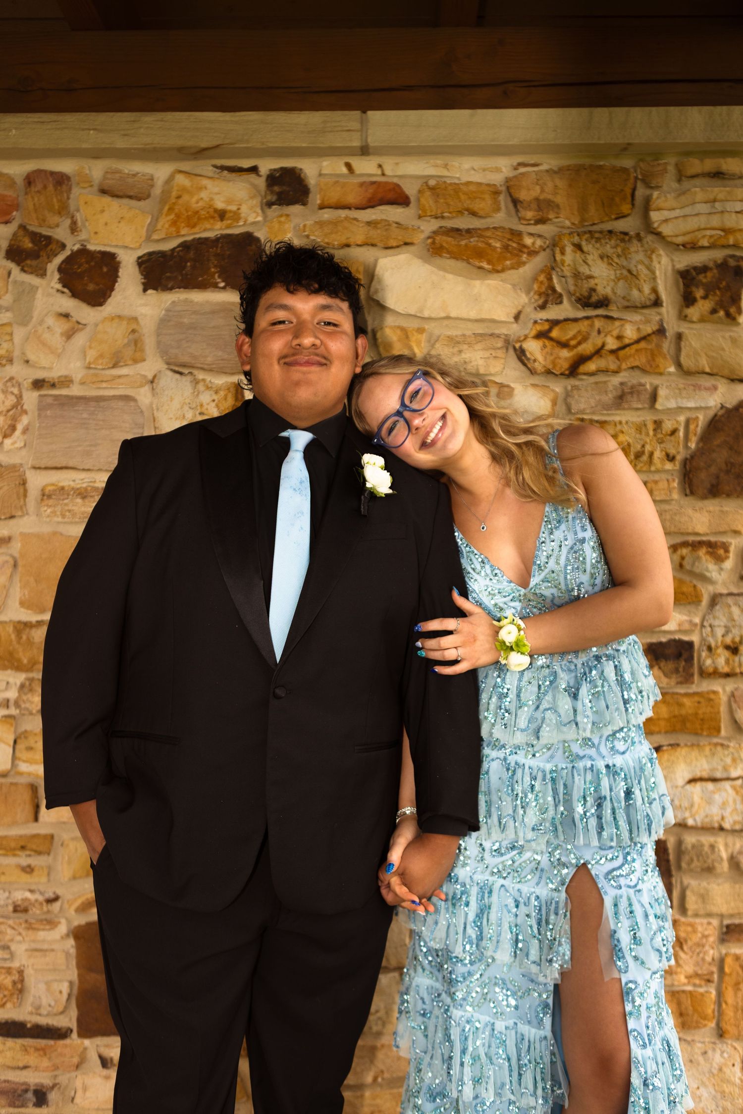 Formal couple in black suit and blue tiered dress posing together against stone wall at special event.