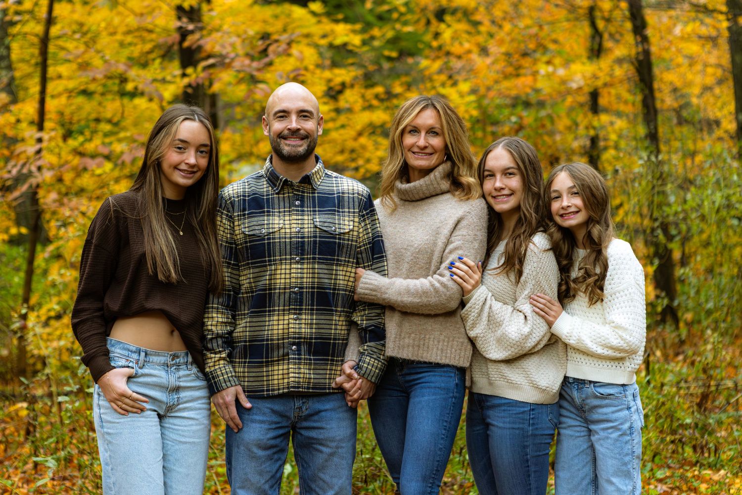 A family group stands together smiling outdoors among vibrant yellow fall foliage in a forest setting.