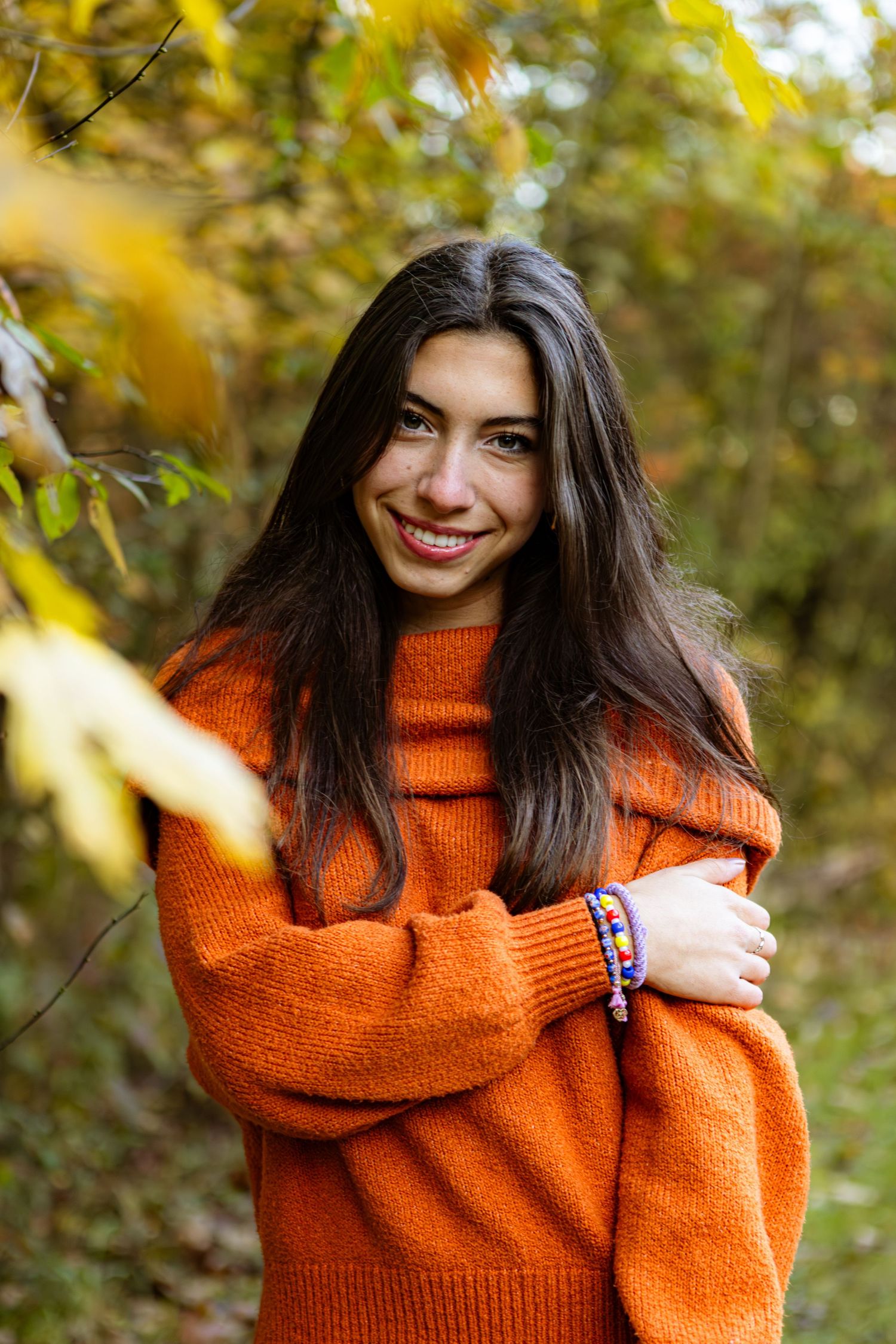 Woman in bright orange sweater smiling outdoors with autumn foliage in background.