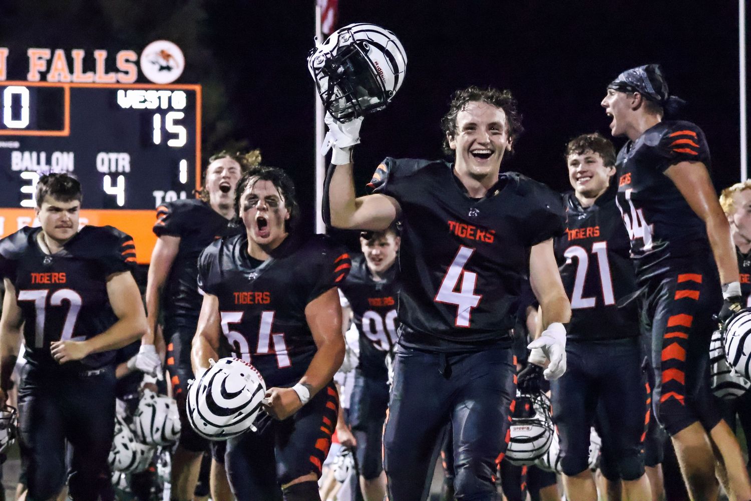 Football team celebrating victory on field at night with players raising helmets.