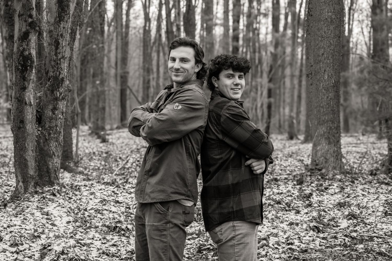 Black and white photo of two people standing back to back in a forest with bare trees during autumn.