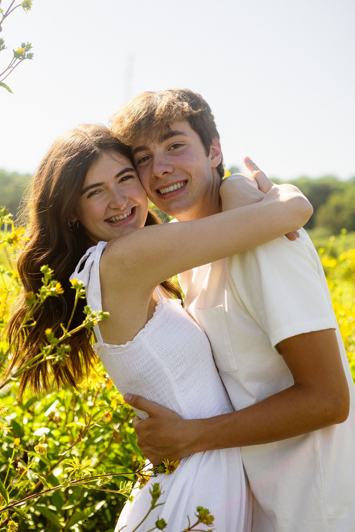 A happy couple embraces outdoors in a sunlit field with yellow flowers on a warm summer day.