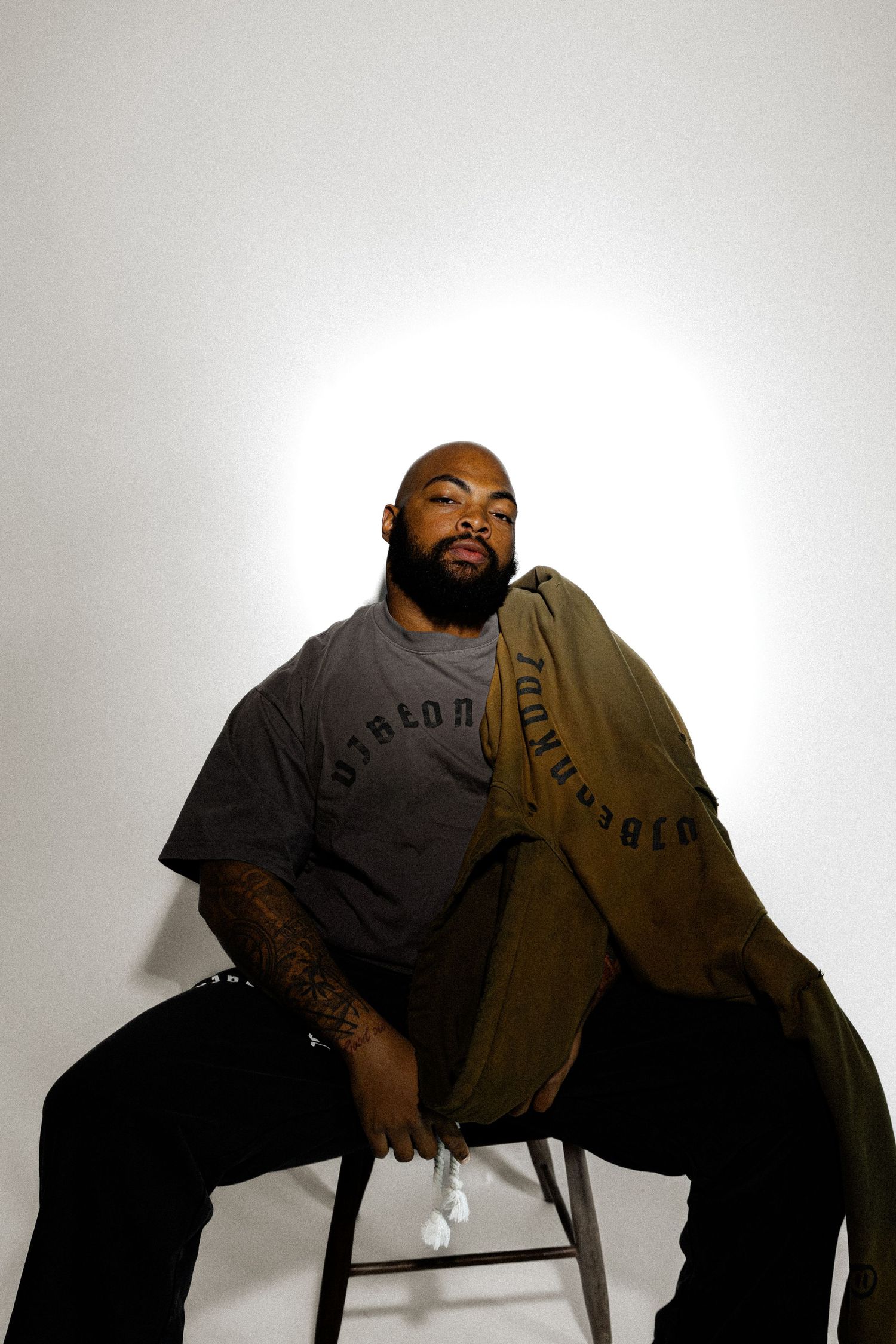 Bearded Black man with tattoos sits on stool holding olive jacket, wearing gray graphic tee and black pants against white backdrop.