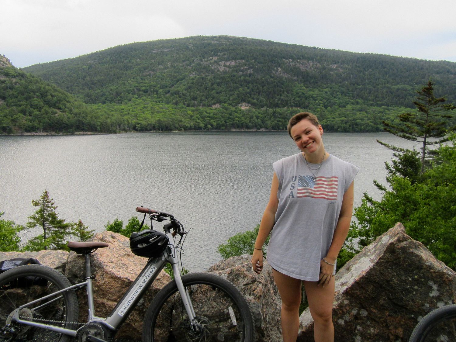 Young person smiling beside a bicycle with a scenic lake and green mountain backdrop on a cloudy day.