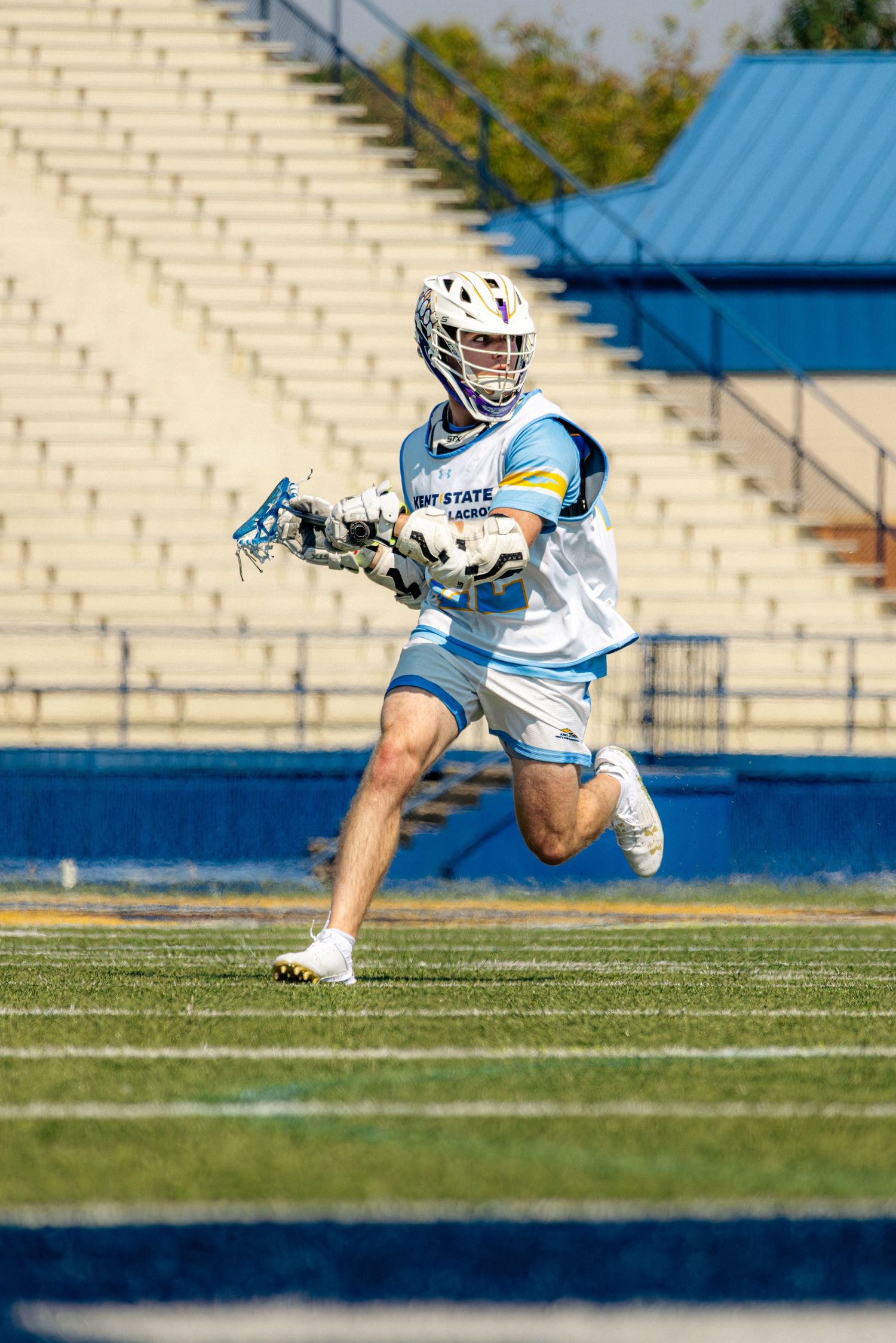 Lacrosse player in white and blue uniform runs across field while carrying stick during daytime game.