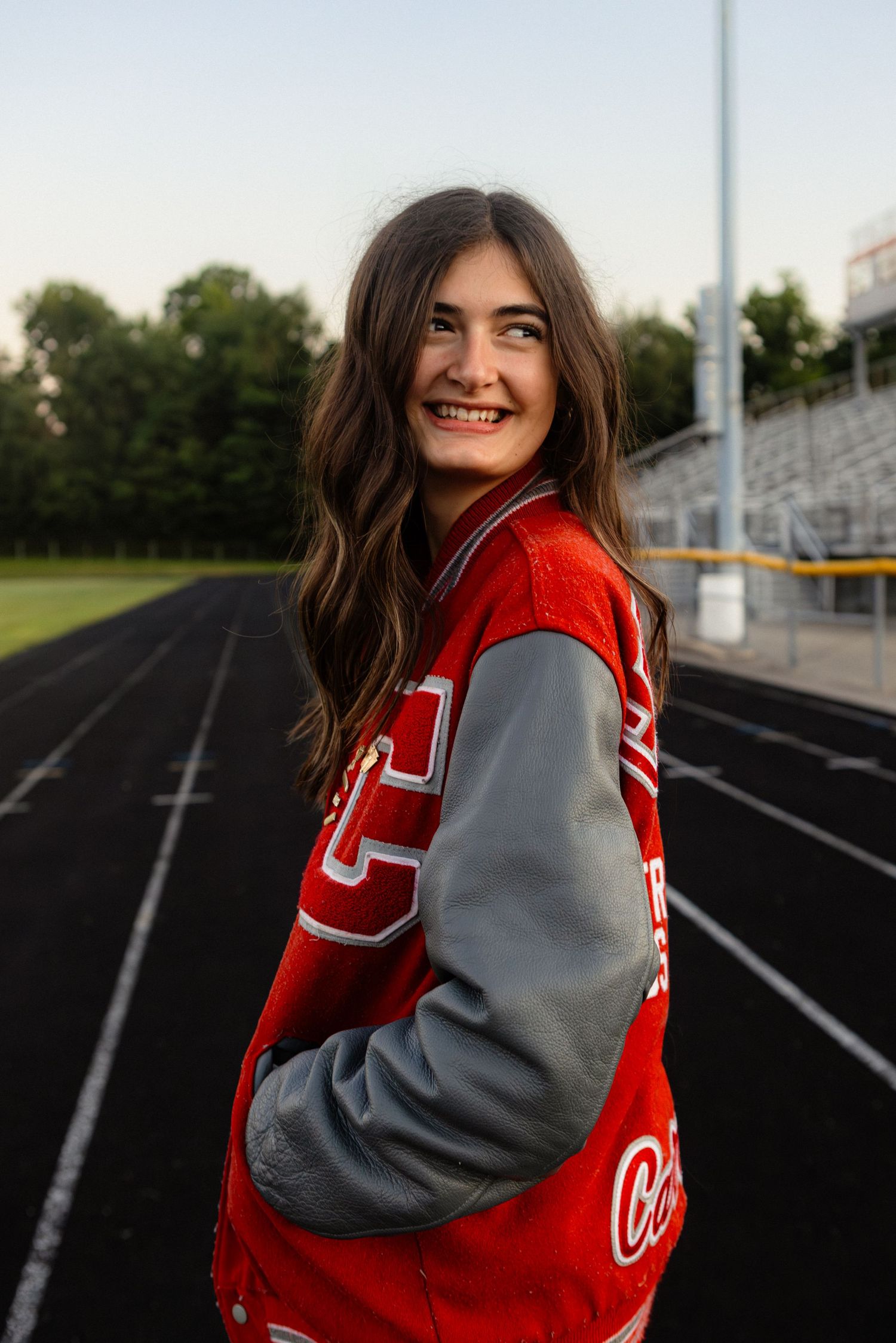 Athlete in red and gray varsity jacket smiles while standing on stadium track during golden hour.