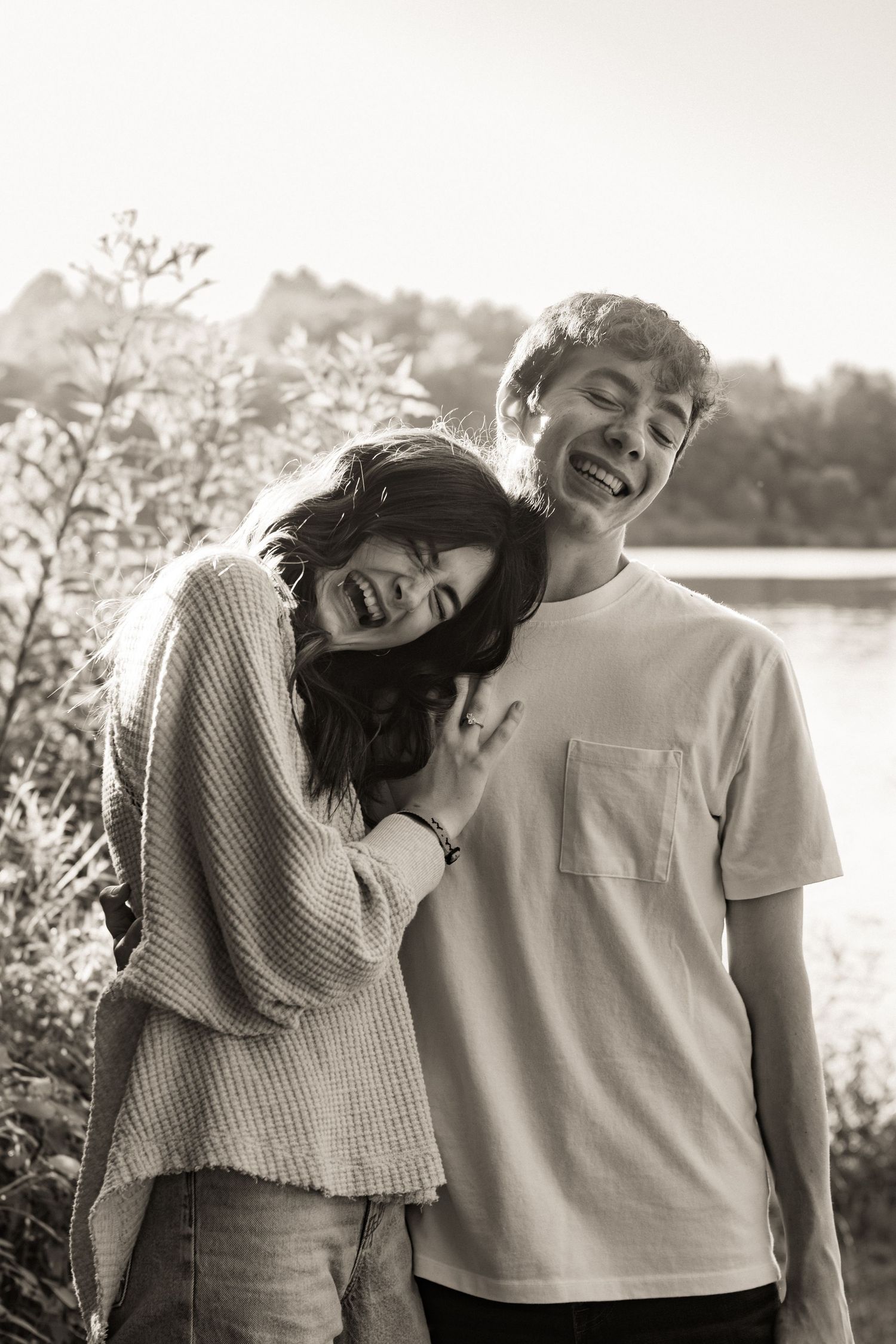 A black and white outdoor portrait of a happy couple laughing and embracing by a lake on a sunny day.