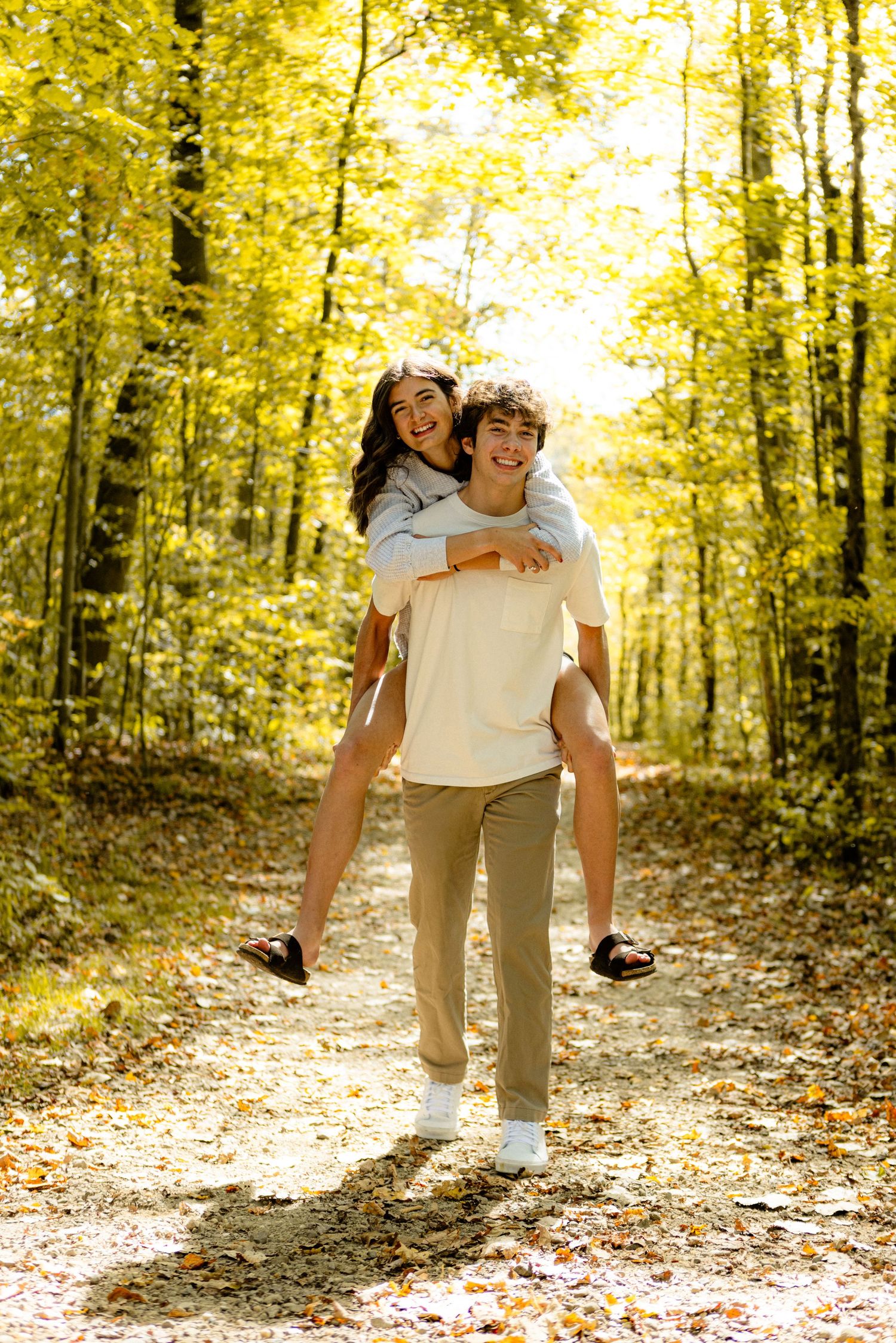 A couple playfully walks through a sunlit autumn forest path covered in fallen yellow leaves.