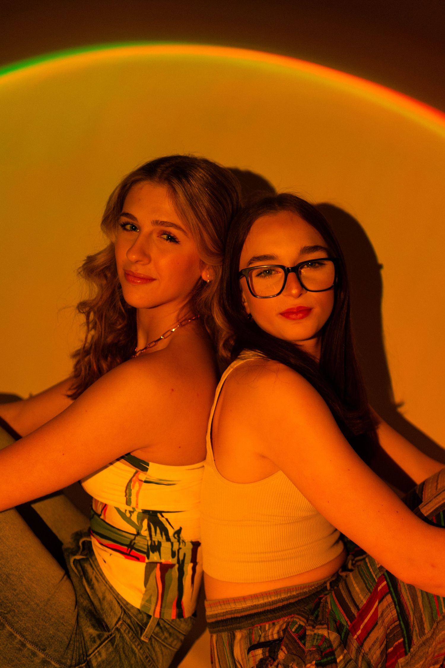 Two friends pose back-to-back in a stylish photoshoot with warm orange lighting and casual crop top outfits.
