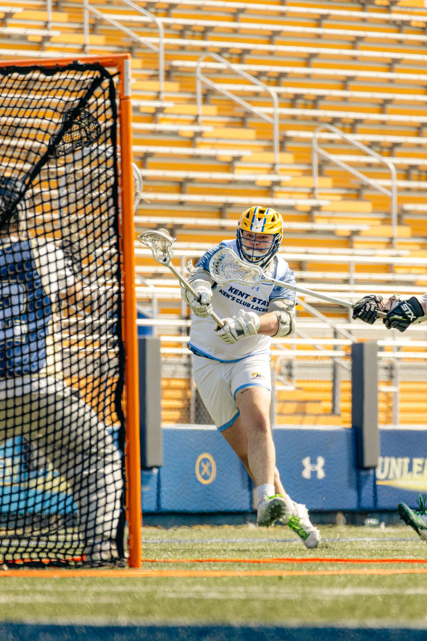 A lacrosse goalie in white uniform makes a diving save attempt in front of the goal net during a match.