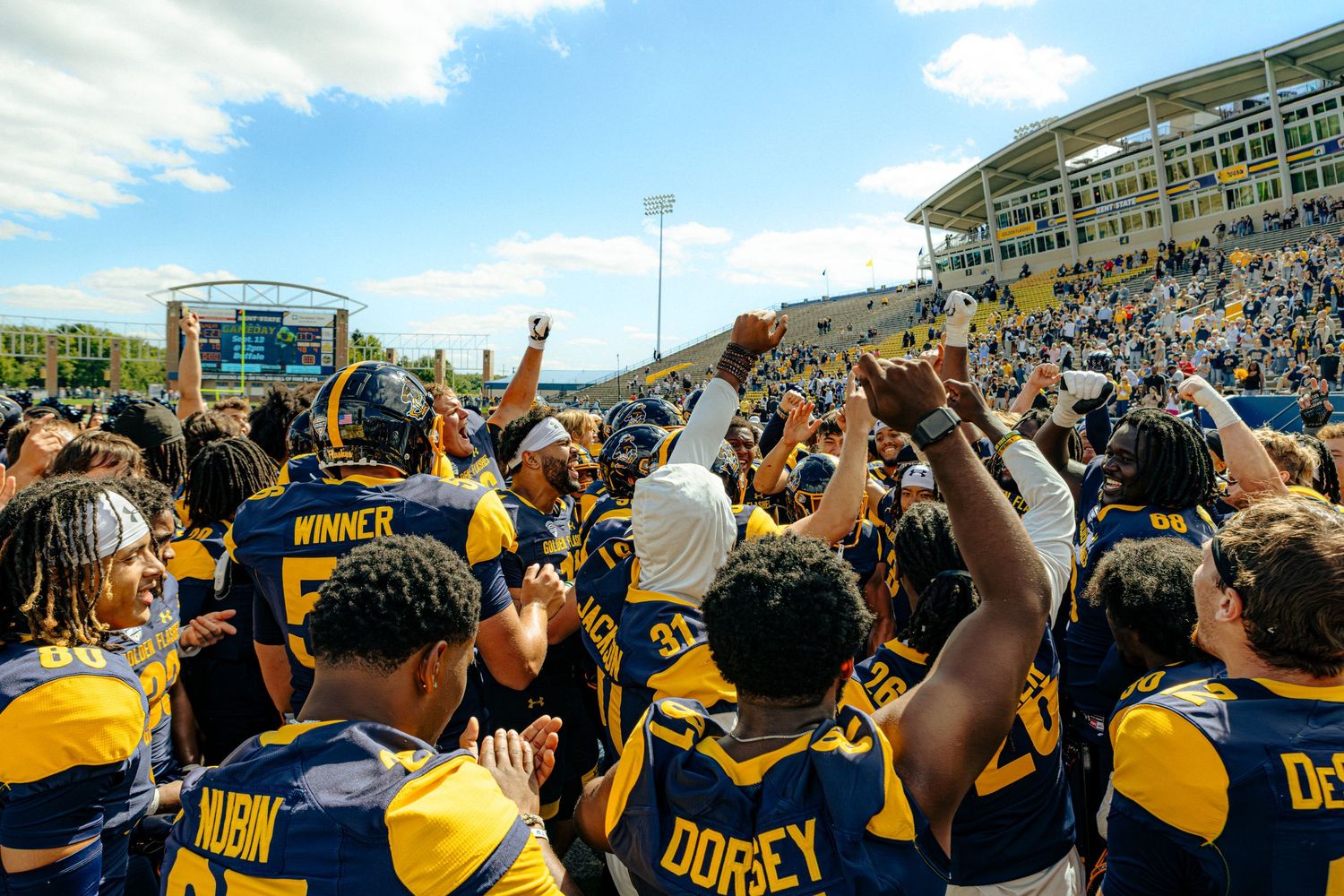 Panoramic view of a crowded football stadium with fans in navy and yellow team colors celebrating with raised arms.