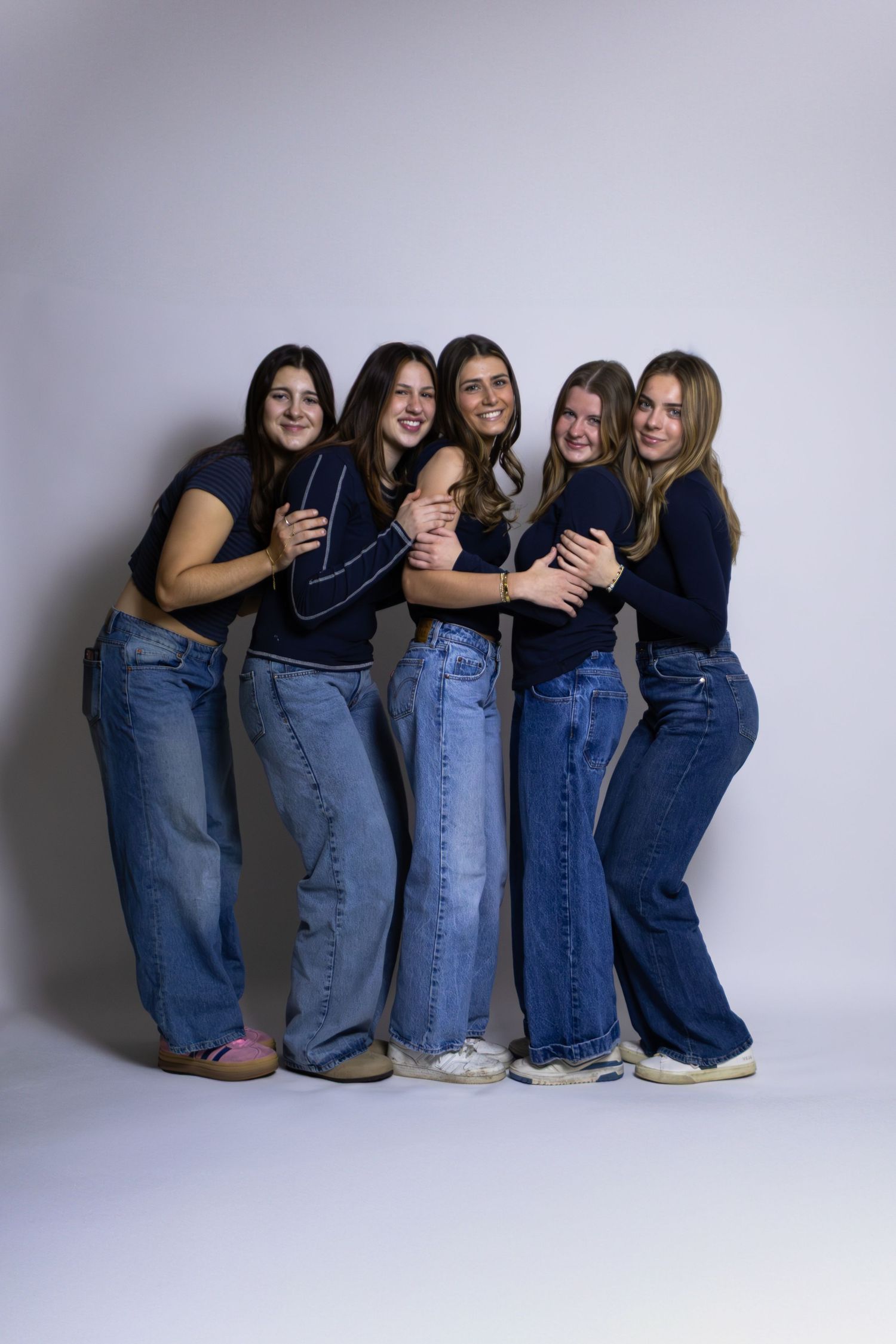 Group of five people in matching blue jeans and navy tops sitting in a row against a white wall.