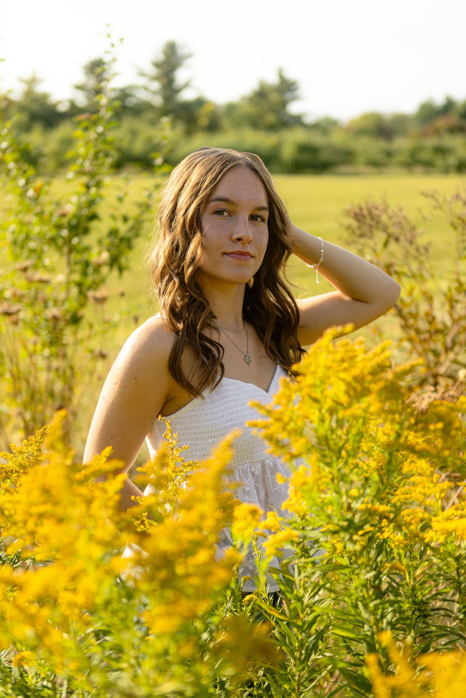 A person in a white top poses thoughtfully among vibrant yellow wildflowers in a sunny summer meadow.