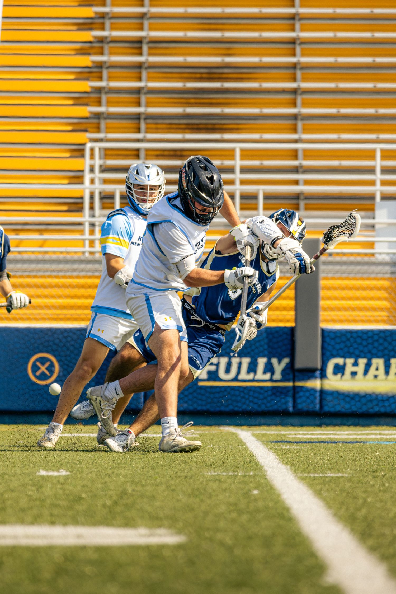 Lacrosse players battle for possession of the ball on a green turf field near the sideline during a match.
